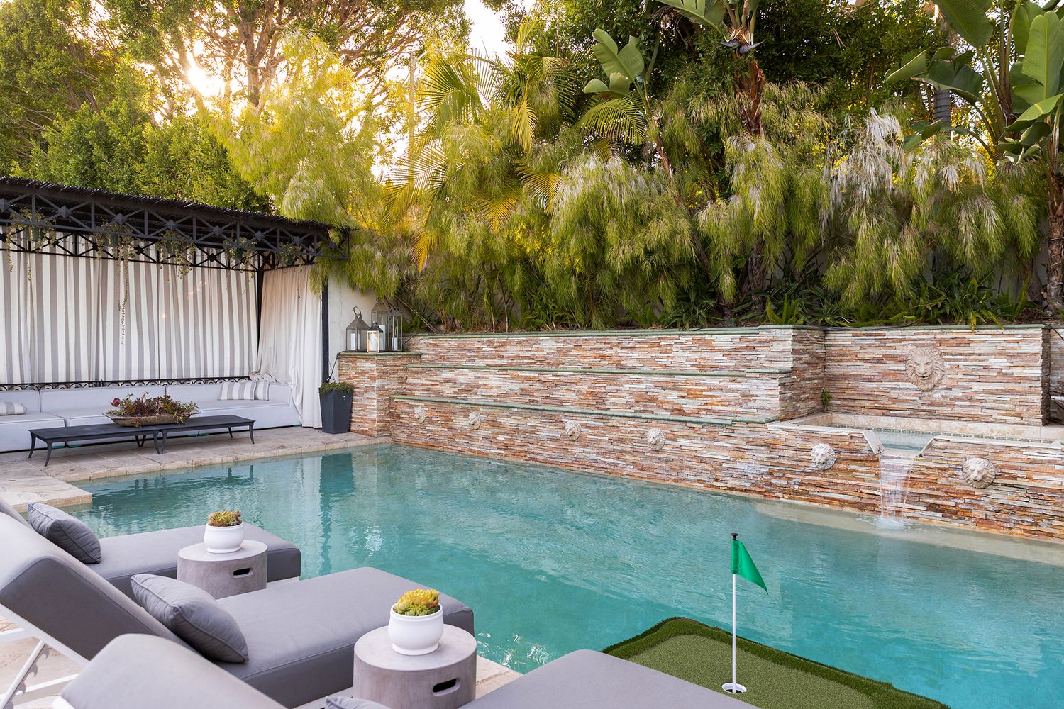 Luxury backyard pool area with stone walls, modern lounge furniture, and lush greenery creates a serene outdoor oasis.