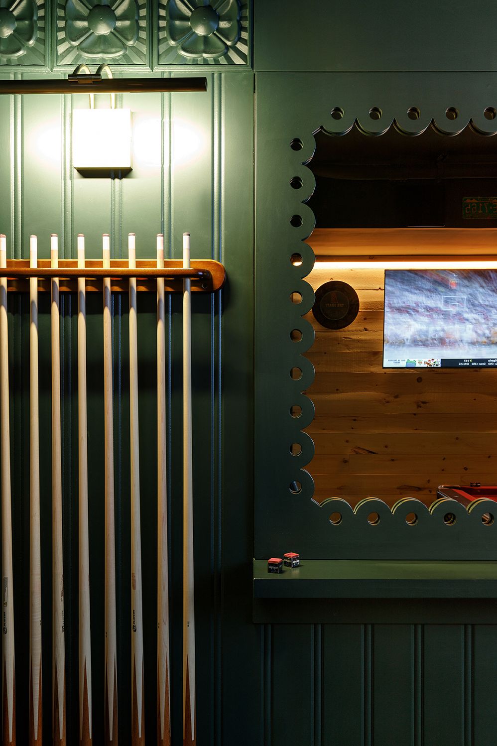 Green-painted billiard room wall with cue rack, scalloped trim, and backlit TV display in a stylish game room.