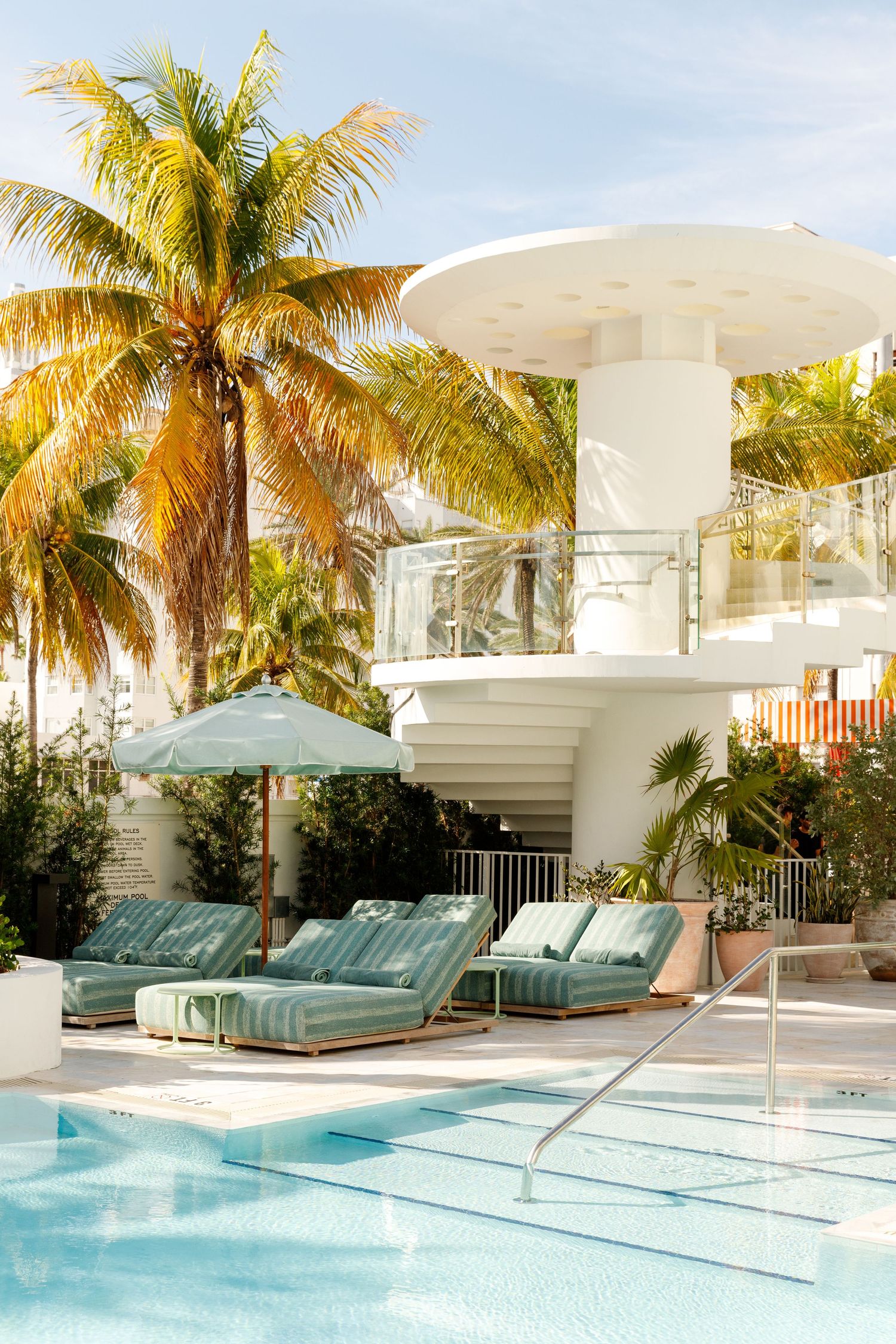 Luxury hotel pool with teal lounge chairs, umbrella, palm trees, and white Art Deco architecture in Miami Beach.