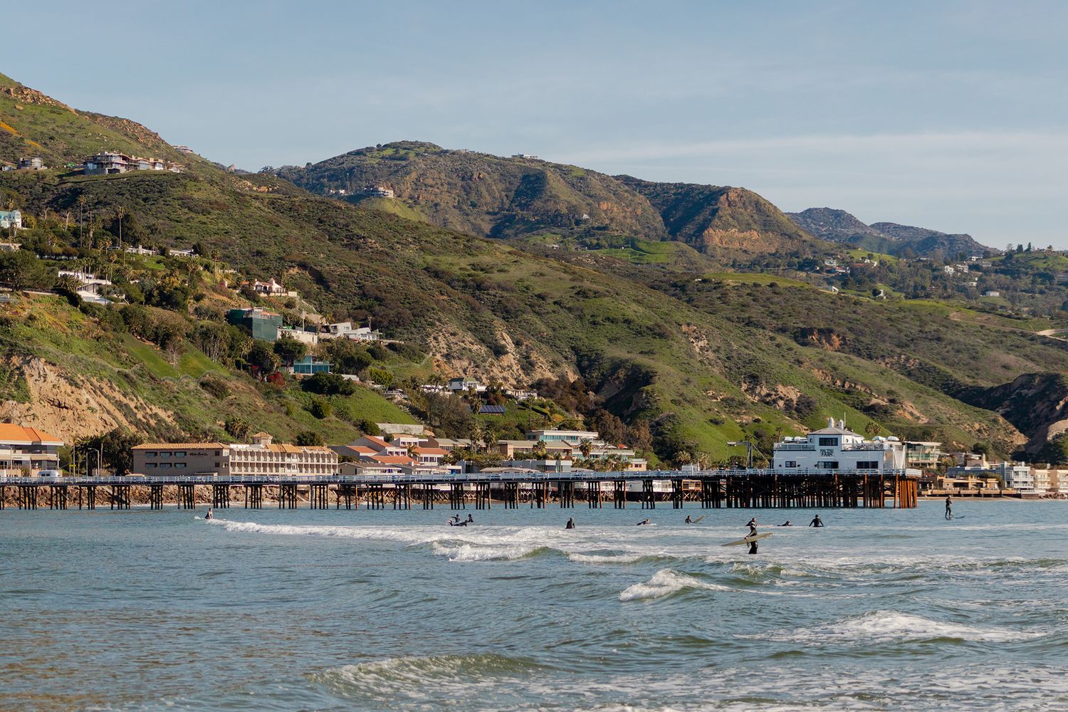 Scenic Malibu Pier stretching over calm ocean waters with surfers, green hills, and coastal buildings in the background.