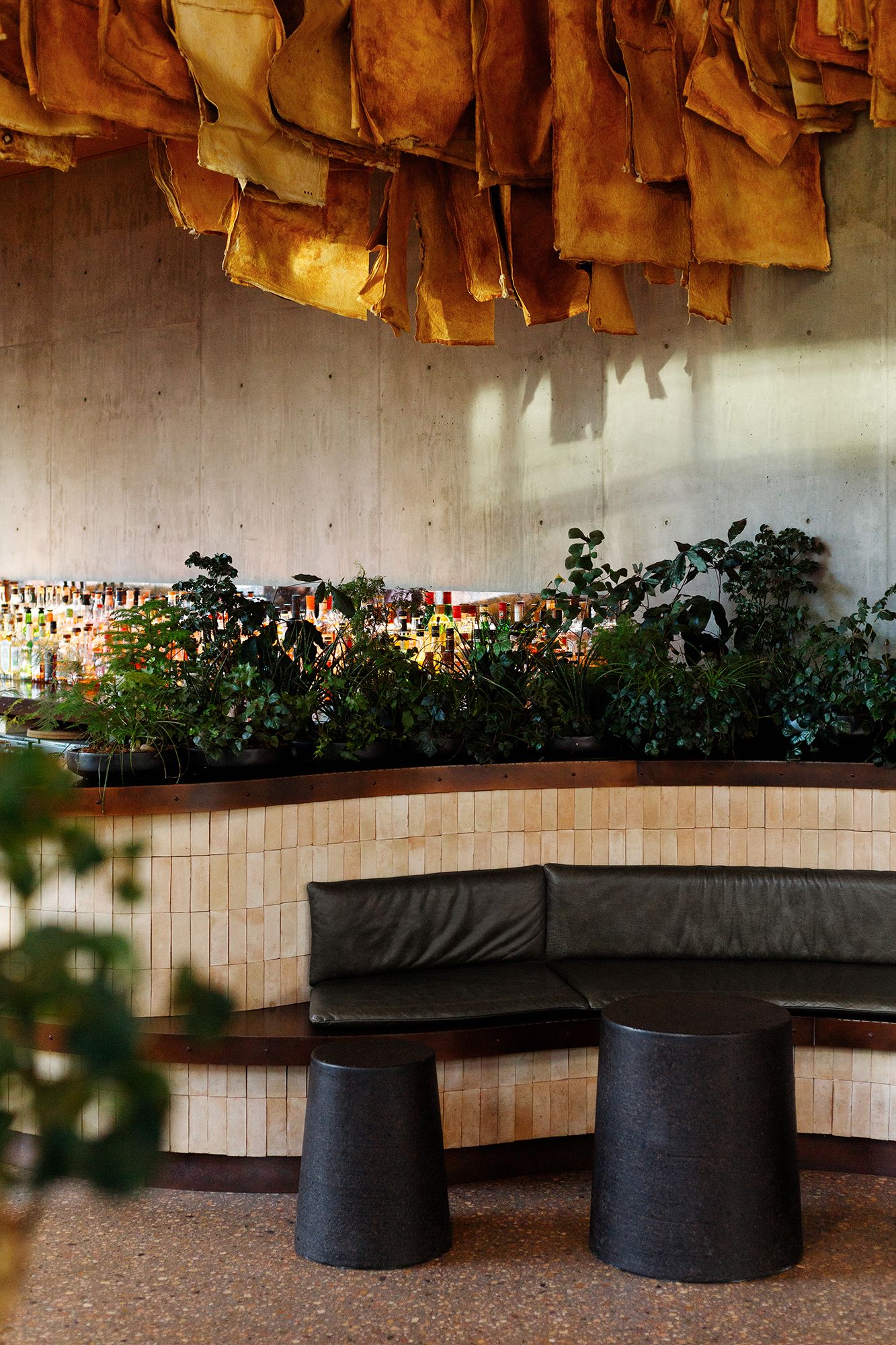 Modern restaurant interior with curved bench seating, black cylindrical stools, plants, and amber-colored decor hanging above.
