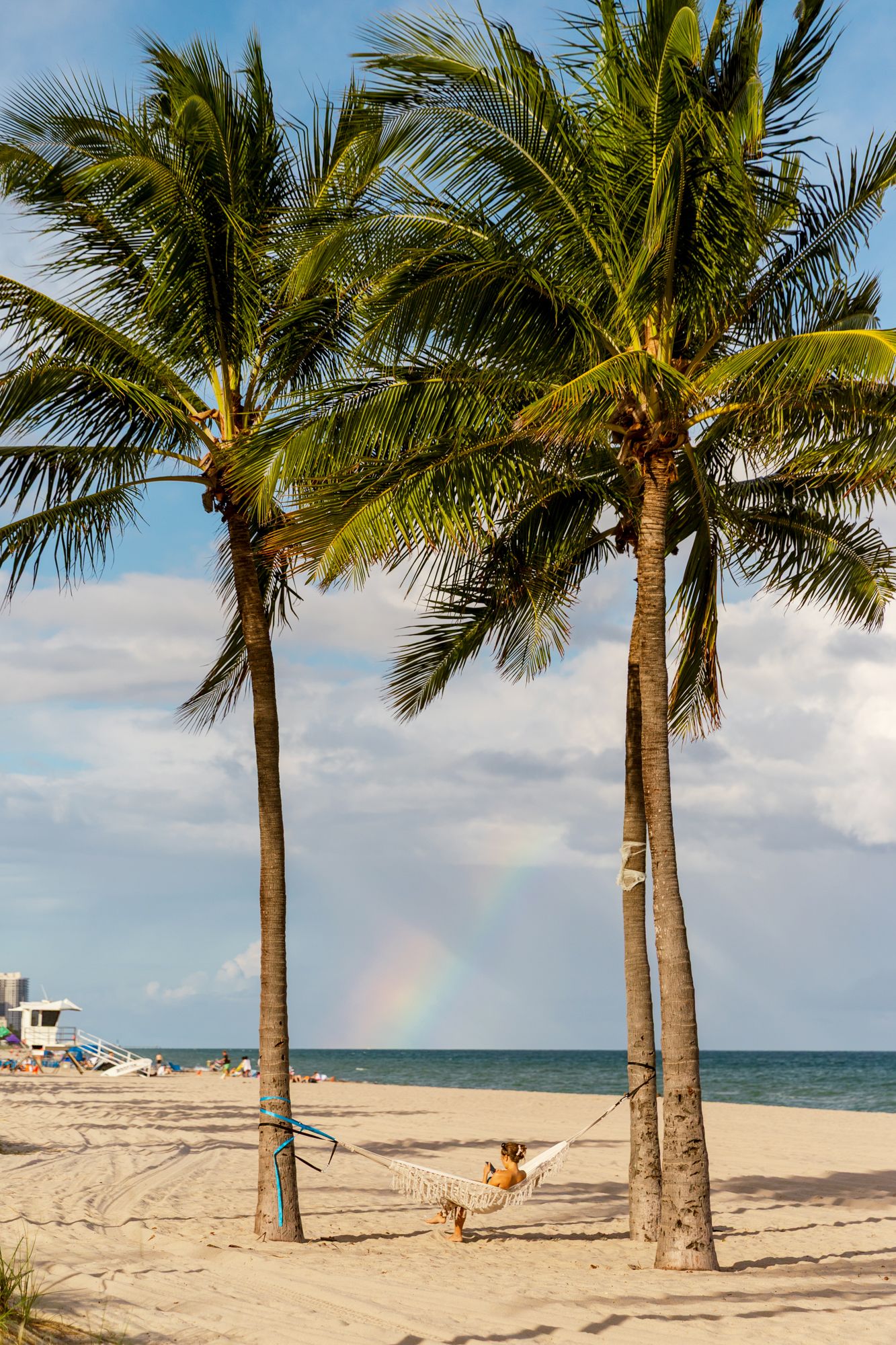 Palm trees line a pristine white sand beach with a rainbow visible in the tropical sky.