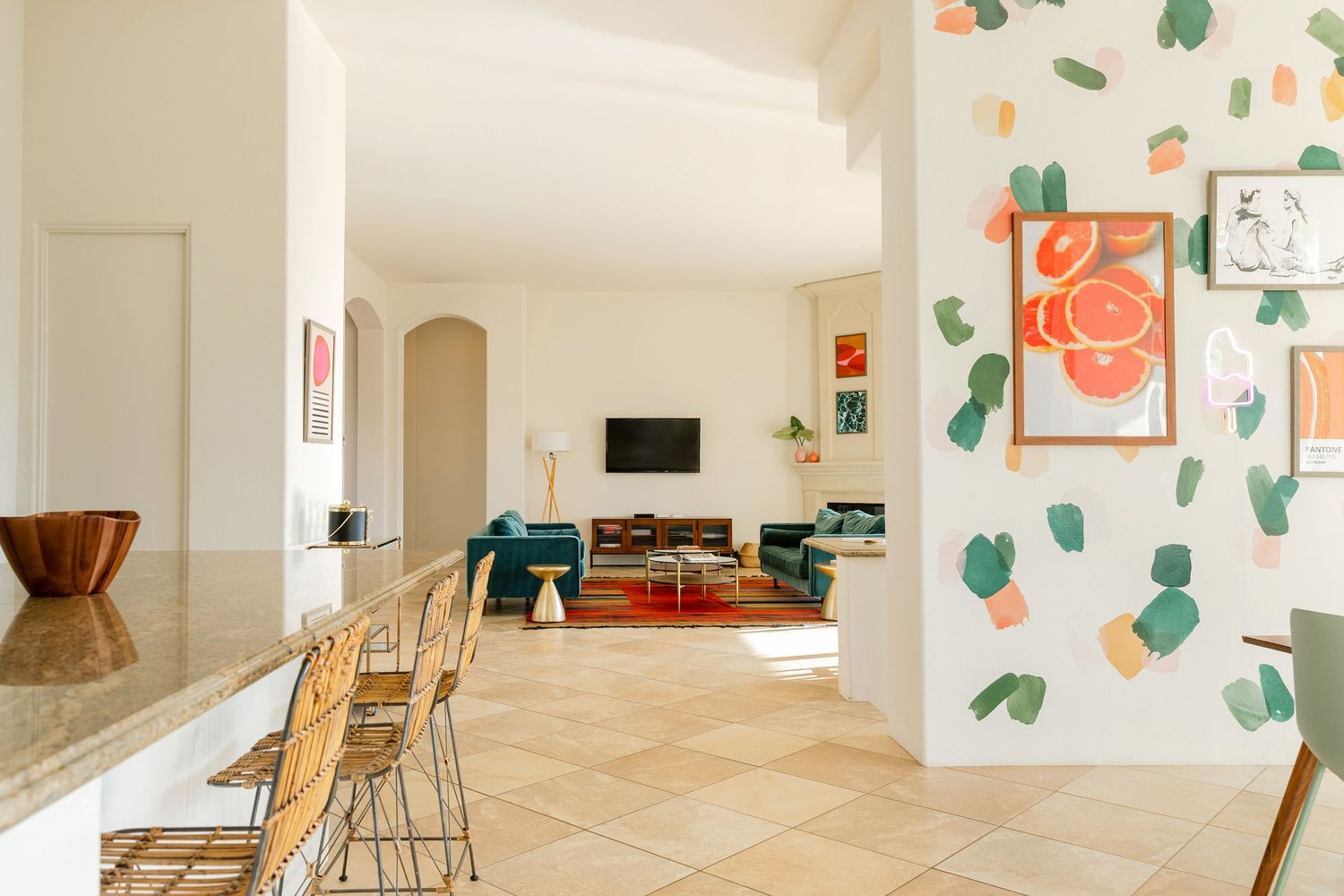 Modern open-plan living space with colorful abstract wall art, teal sofa, rattan chairs, and tile flooring.