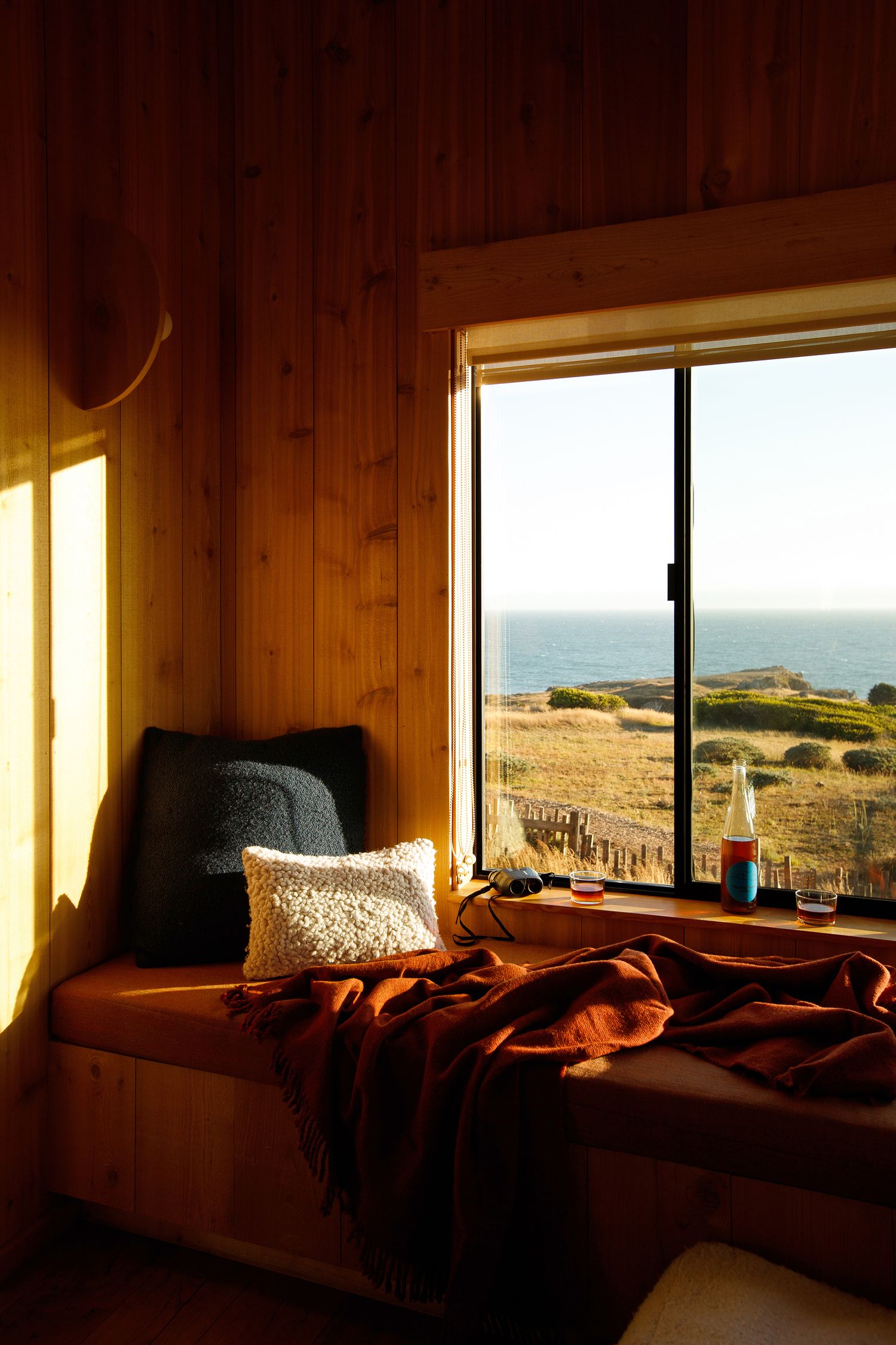 Cozy wooden cabin window seat with orange throw blanket and ocean view at golden hour sunset.