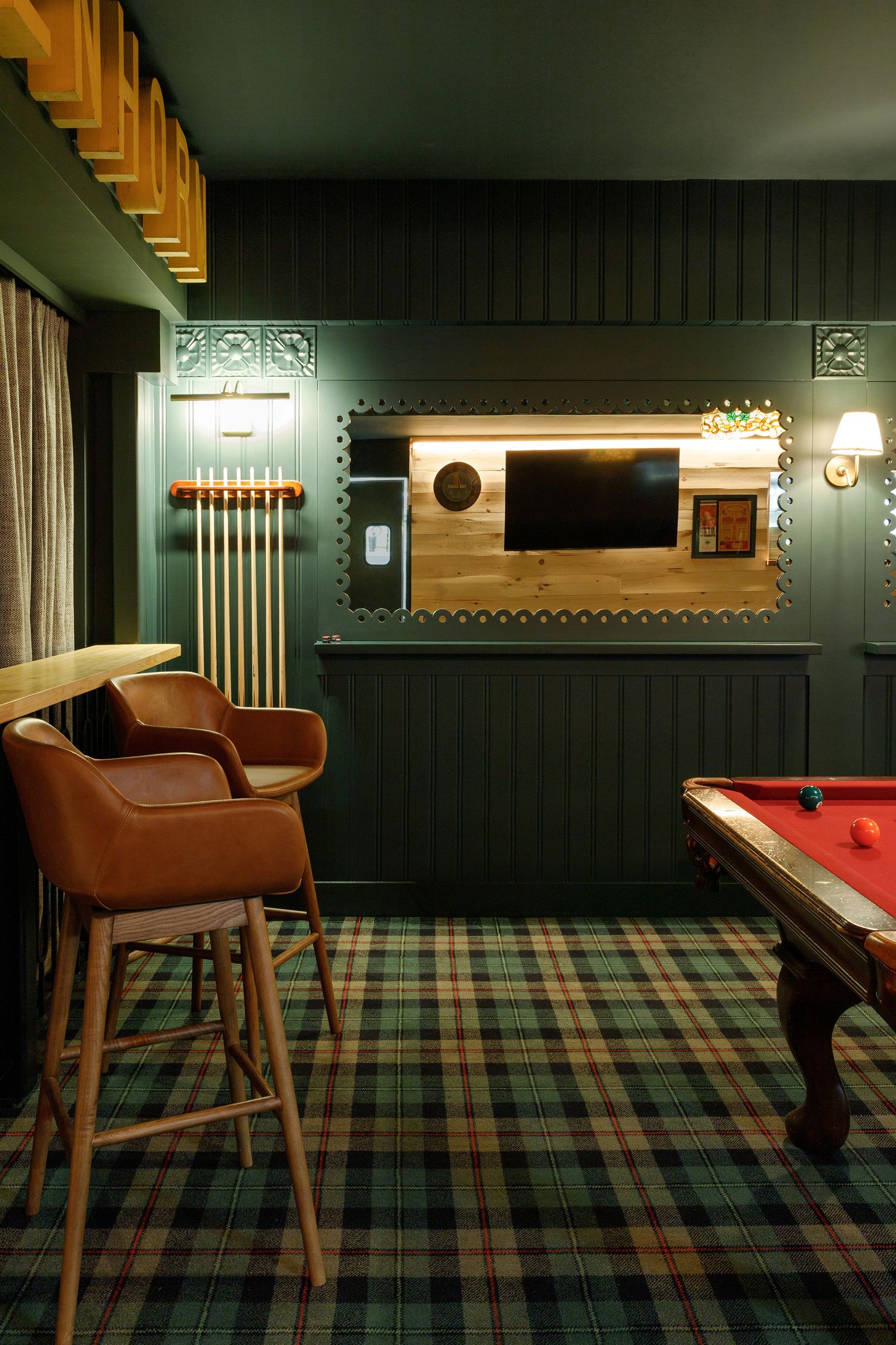 Cozy game room with green plaid carpet, leather bar stools, pool table, cue rack, and wall-mounted TV in dark green paneling.