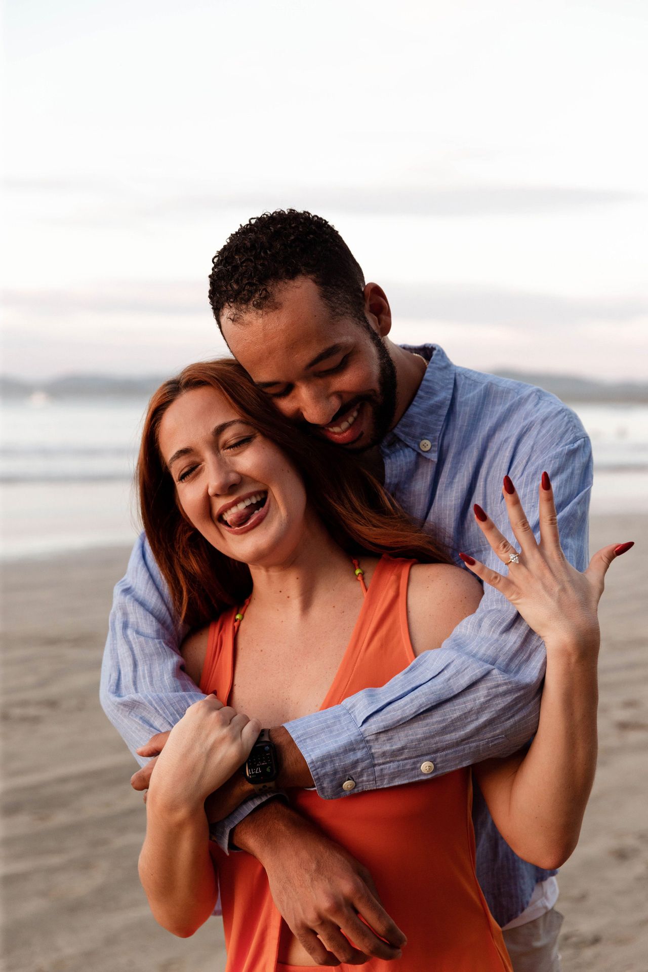 A joyful couple embraces and laughs together on a cloudy beach at sunset.