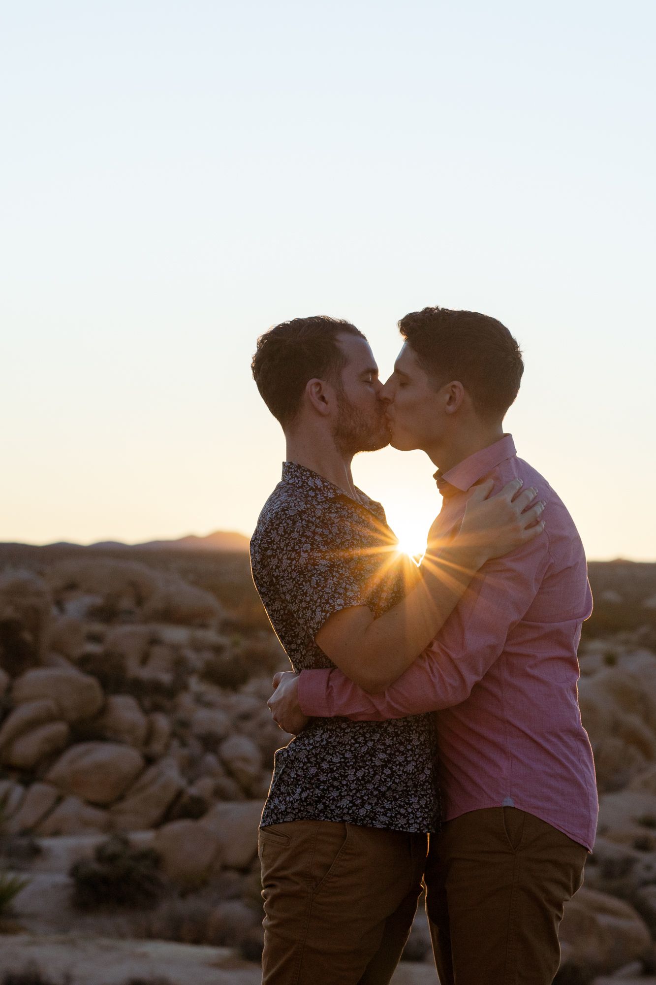 A romantic couple embraces in silhouette against a golden sunset in a desert landscape with rocky terrain.