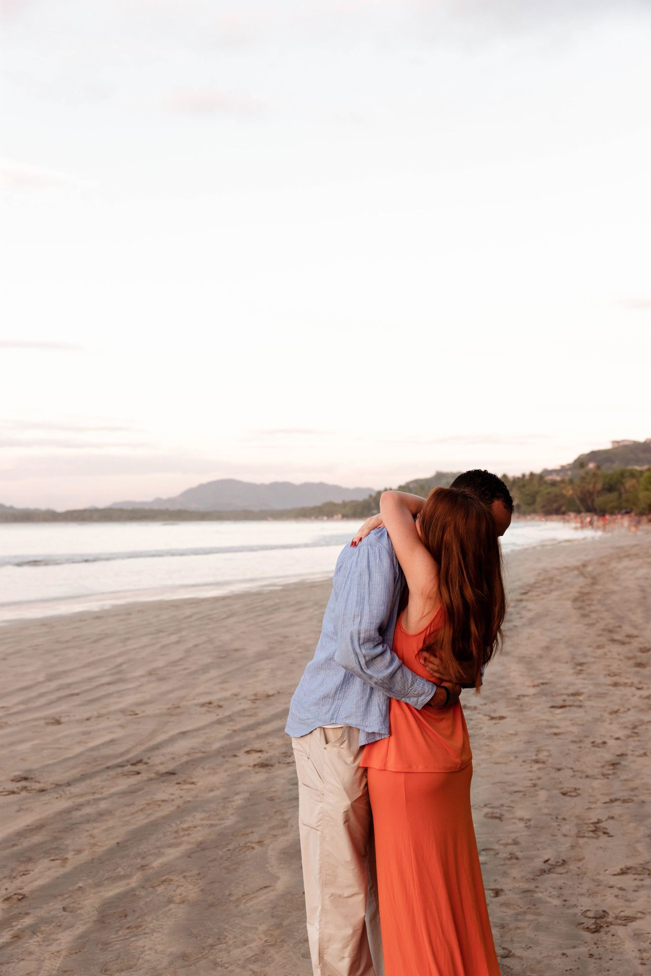 A romantic embrace between a couple in blue shirt and coral dress on a scenic beach at dusk.
