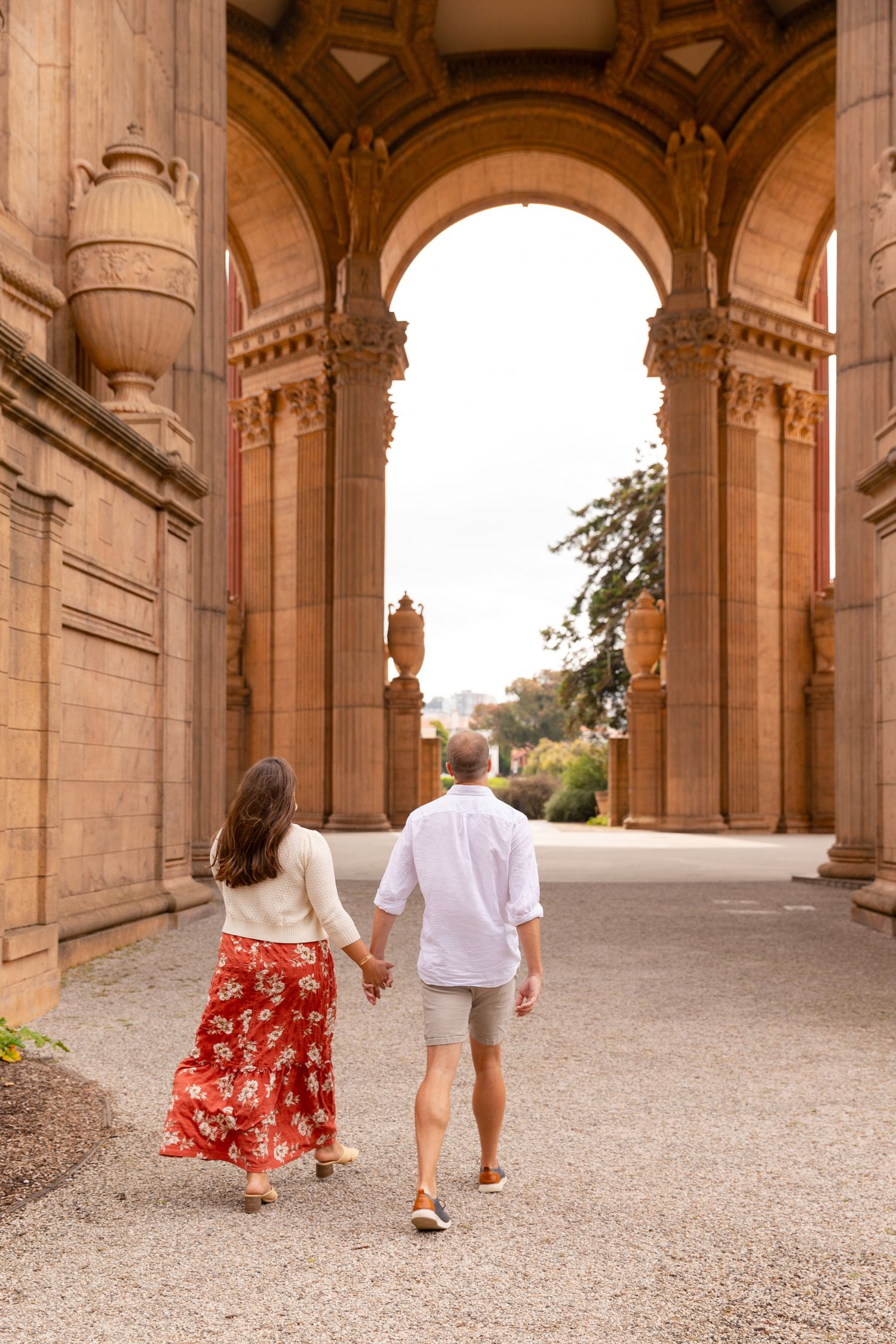 A couple walks hand in hand through a grand stone archway corridor with ornate architectural details.