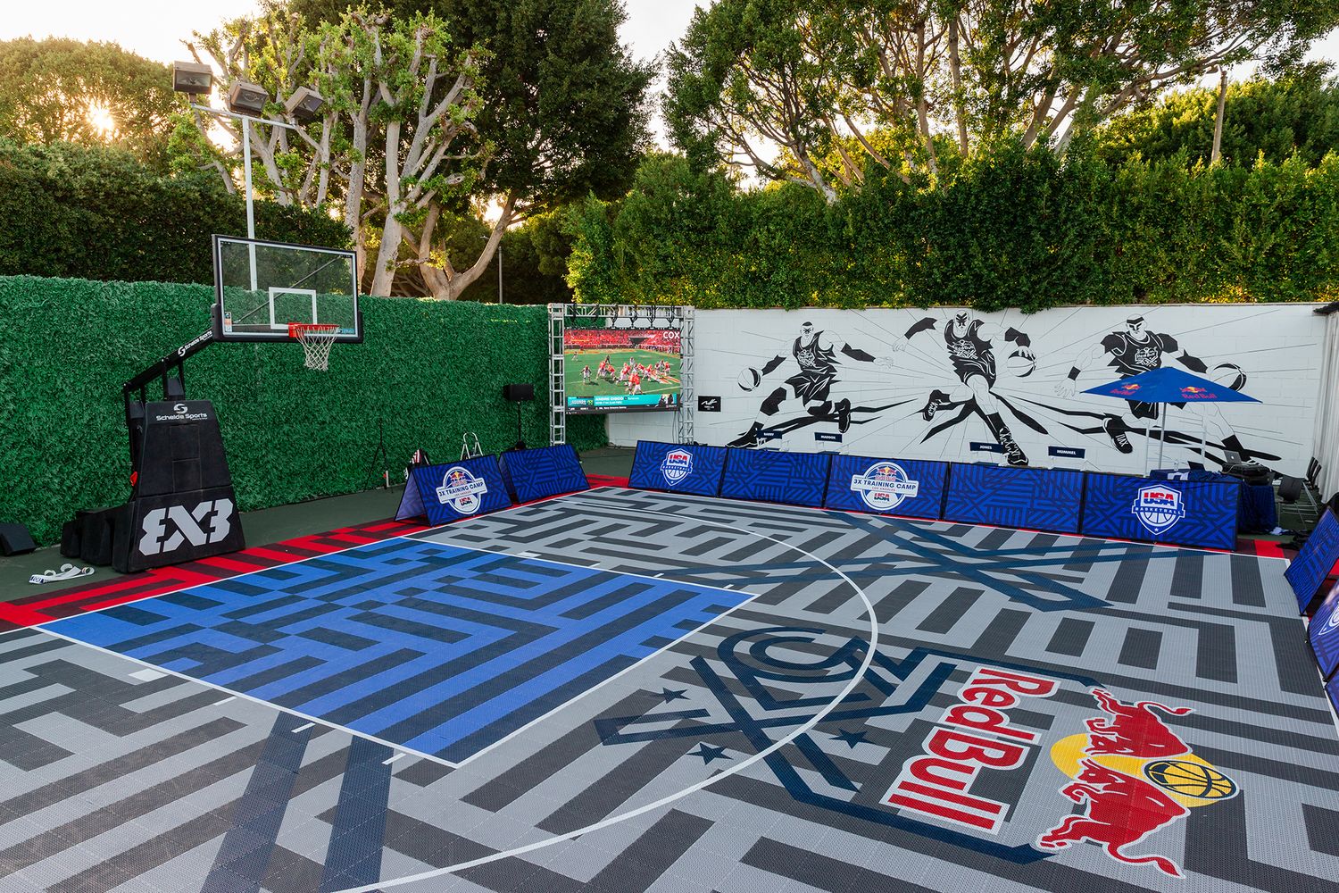 Outdoor basketball court with vibrant geometric patterns and Red Bull branding painted on the surface and green hedge walls.
