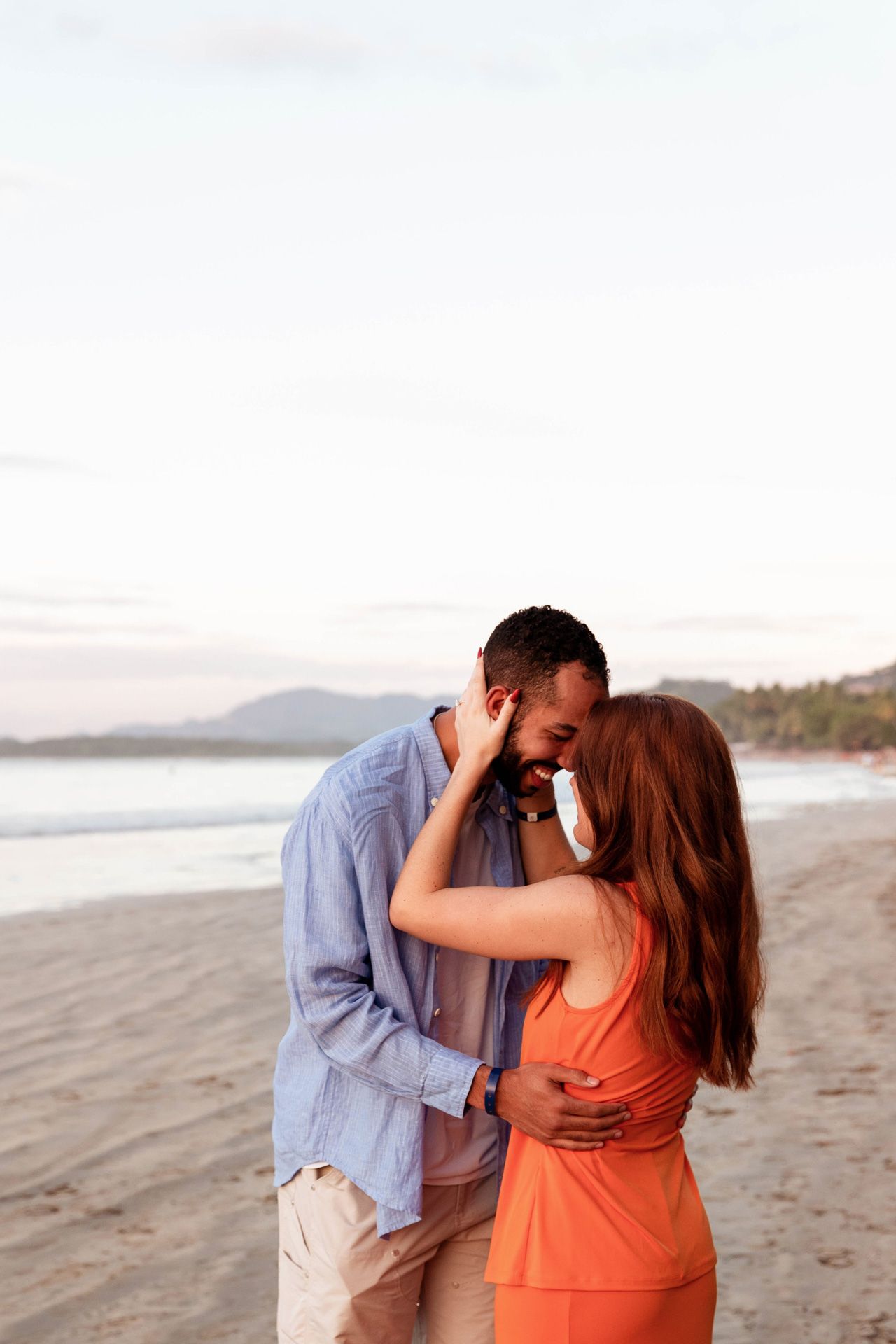 A romantic couple embraces on a scenic beach at sunset, with mountains visible in the background.