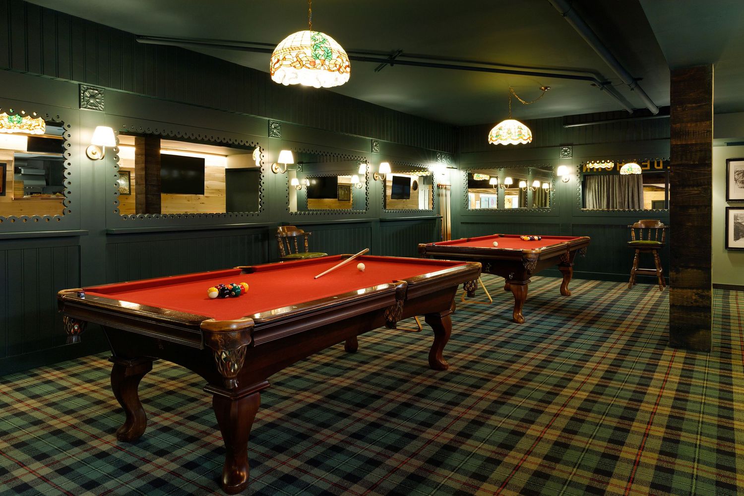 Elegant billiards room with two red felt pool tables, plaid carpet, and warm pendant lighting in a classic wood-paneled setting.