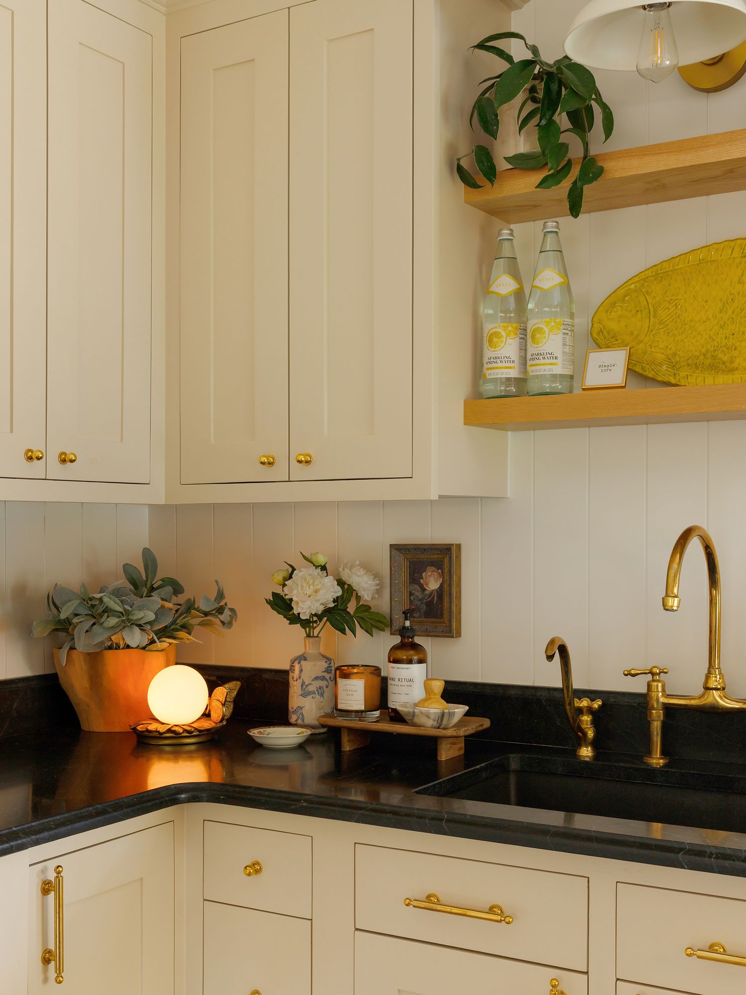 Cream kitchen cabinets with brass hardware, black countertops, open shelves, and a brass faucet sink in a cozy corner.