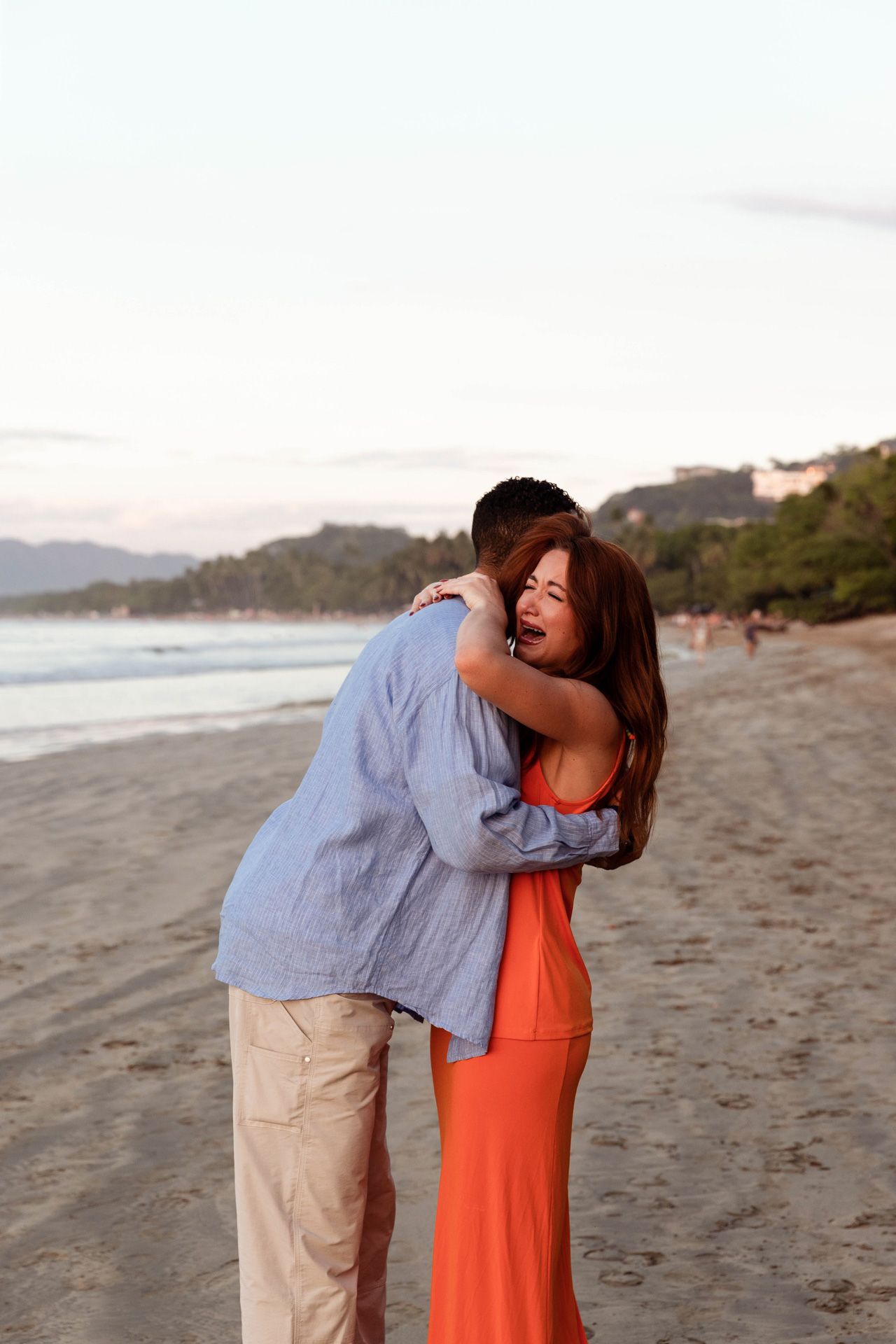 A couple shares a joyful moment together on a beach at sunset wearing orange and light blue outfits.