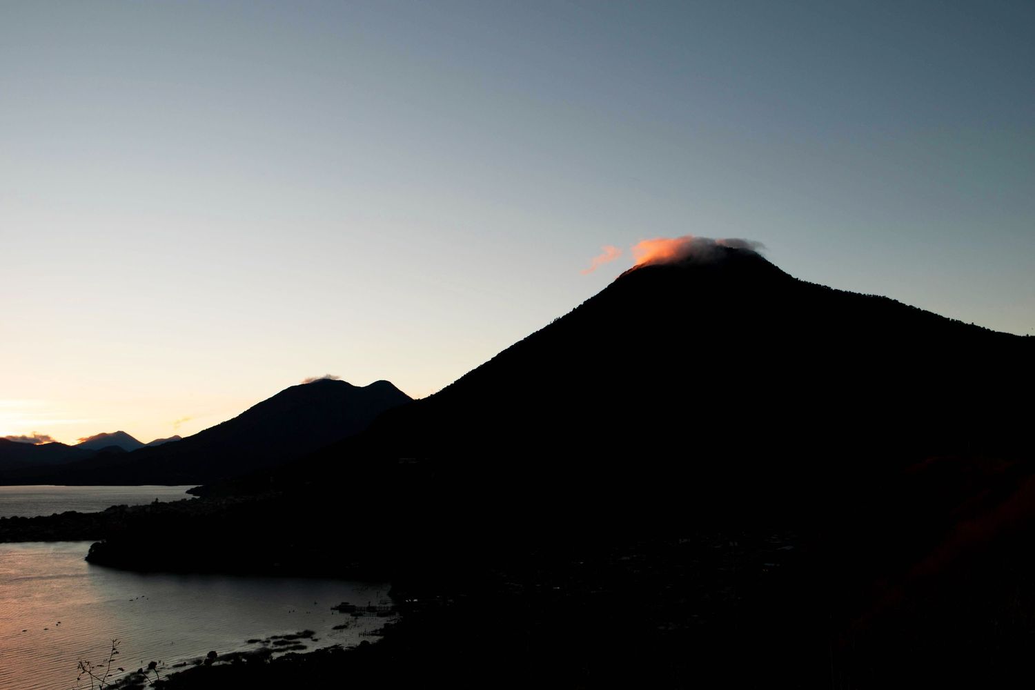 Editorial Travel Photography of Dark silhouette of a volcano in Guatemala, circled by a pink cloud against a fading twilight sky.