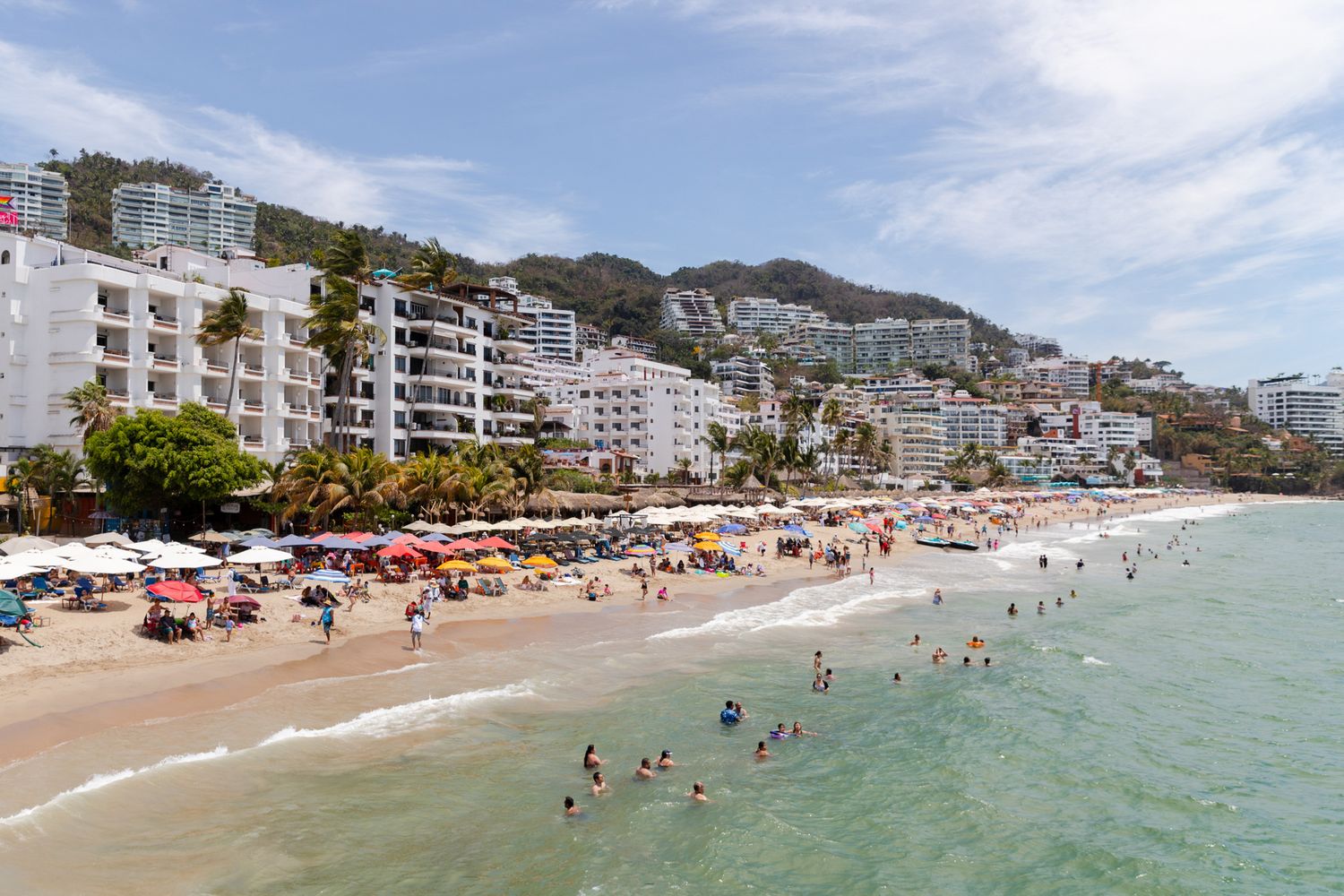 Crowded beachfront with buildings and umbrellas along a turquoise coastline on a sunny day.