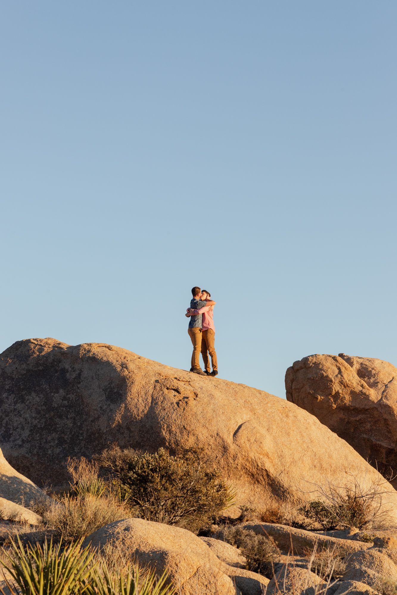 Two people stand atop large desert boulders at sunset in Joshua Tree National Park.