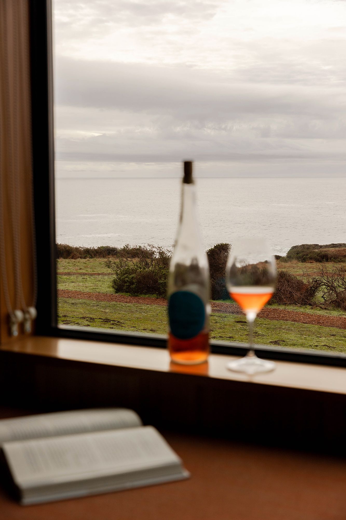 A wine glass sits by a window overlooking a scenic coastal landscape at dusk. Lifestyle, story telling imagery for hotels and resorts.