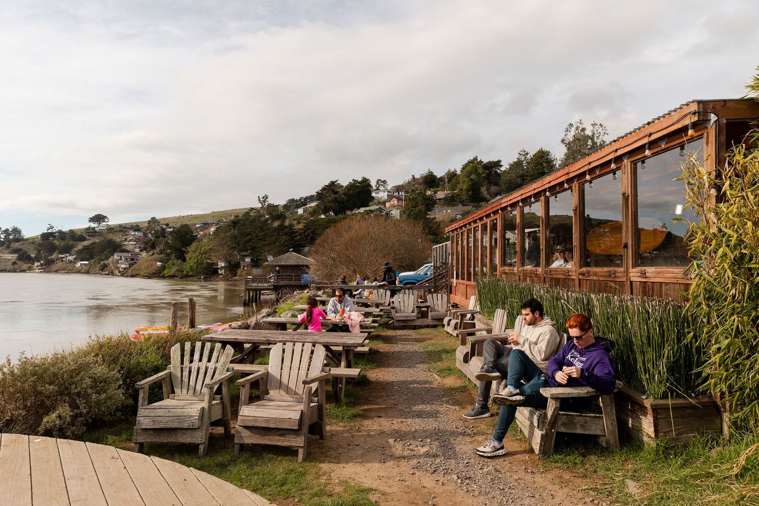 People relaxing on wooden Adirondack chairs along a scenic waterfront path beside a rustic building on a cloudy day.