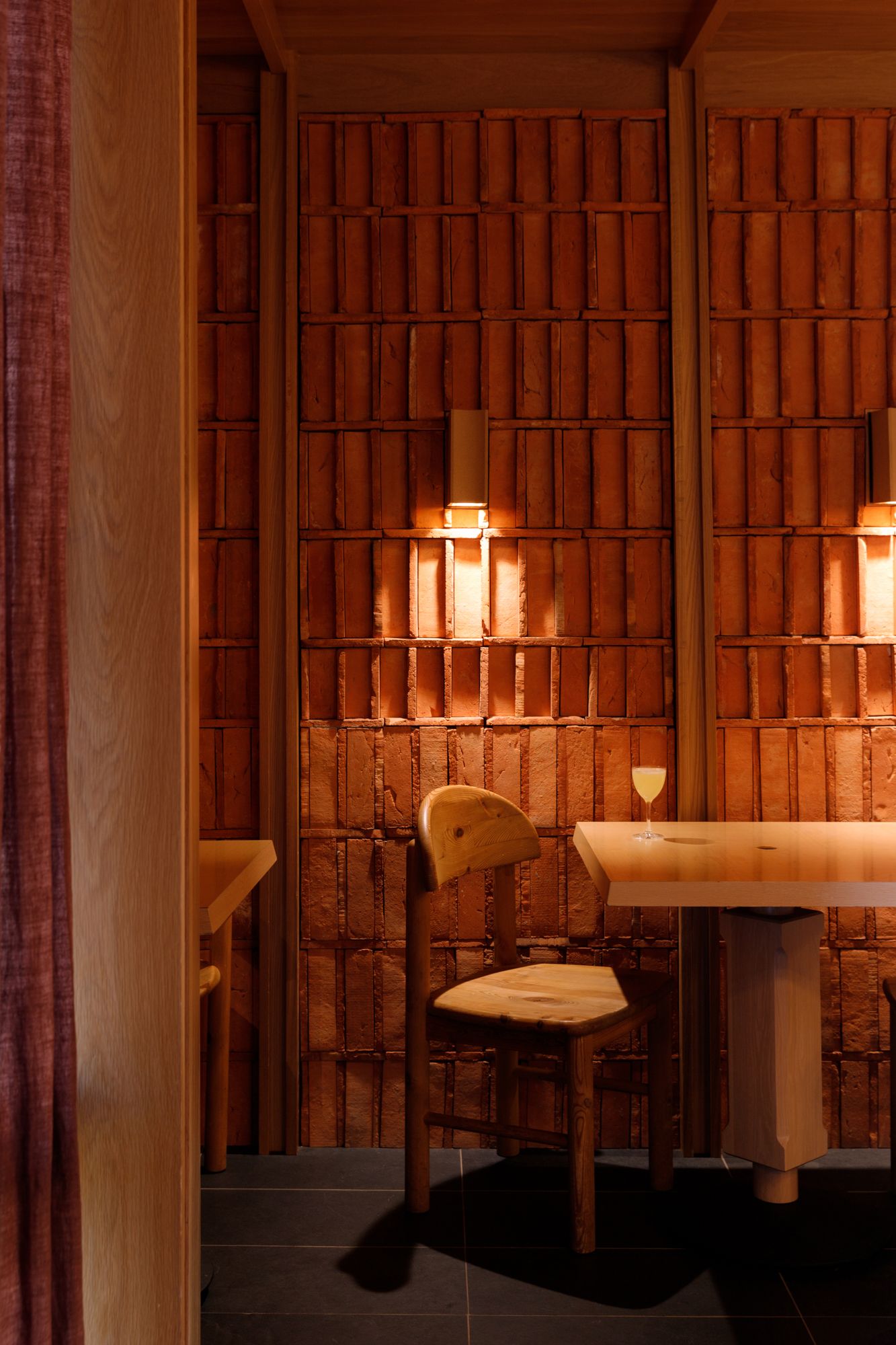 Intimate wooden restaurant booth with warm glowing wall sconces and a single wine glass on a natural wood table.