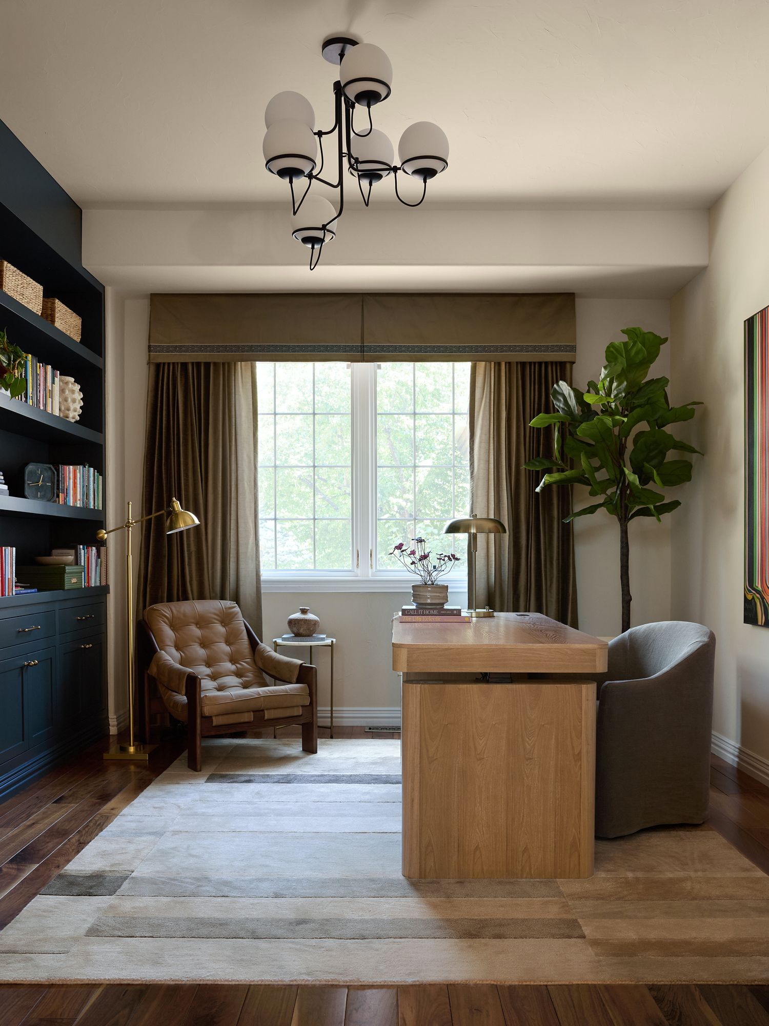 Elegant home office with wood desk, tufted leather chair, dark built-in shelves, fiddle leaf fig tree, and chandelier lighting.