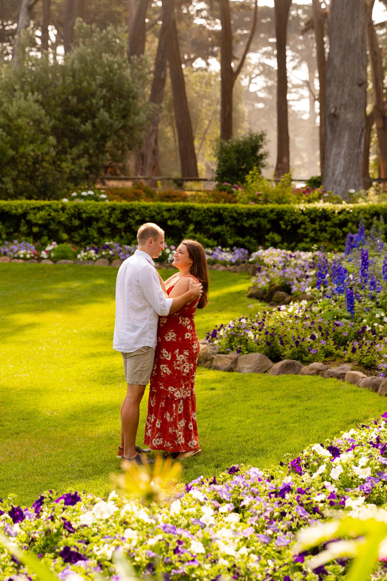 A couple shares a romantic moment in a lush garden filled with vibrant yellow and purple flowers on a sunny day.