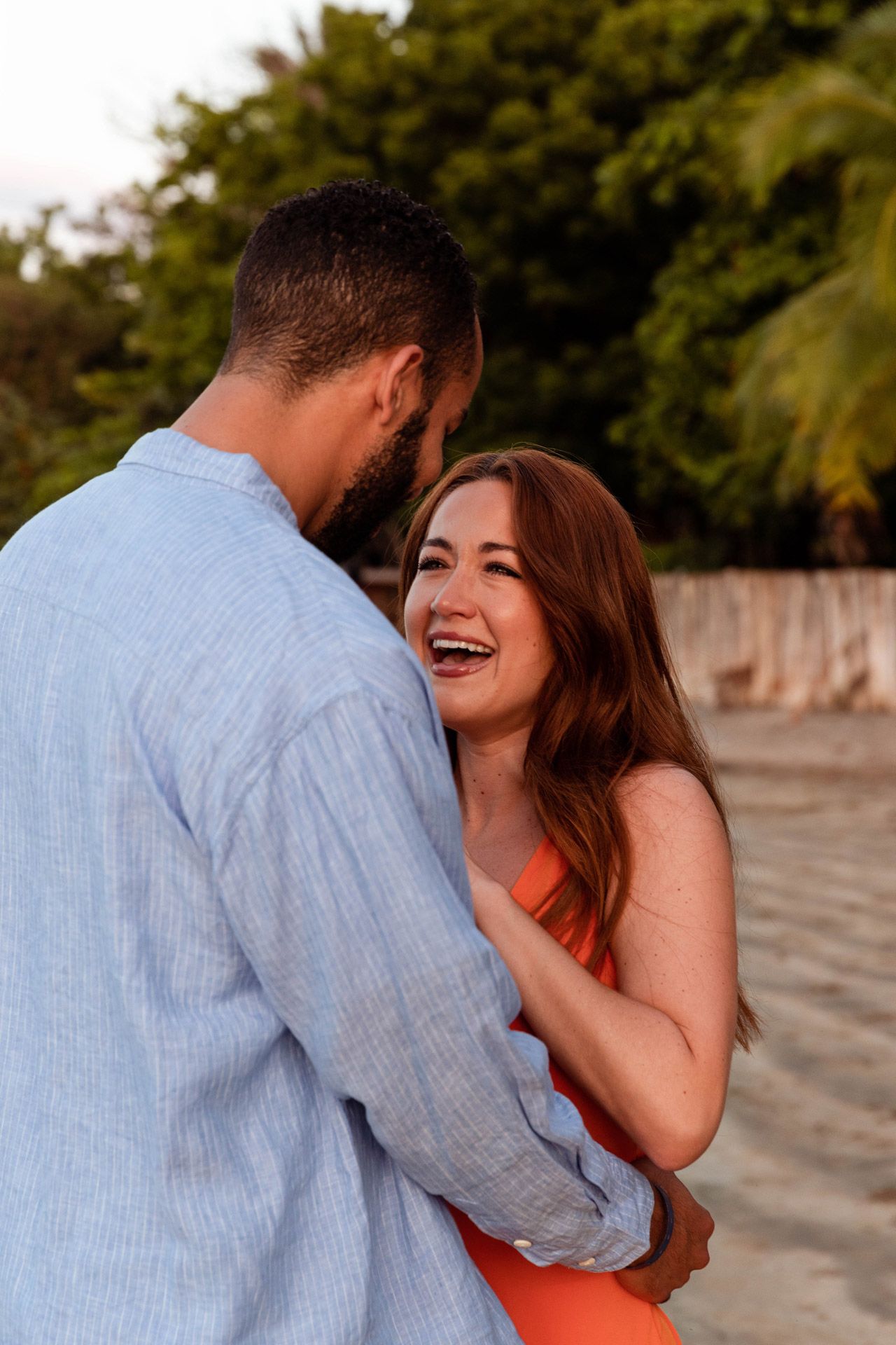 A romantic couple shares a tender moment on the beach with the woman wearing a coral dress and man in blue shirt.