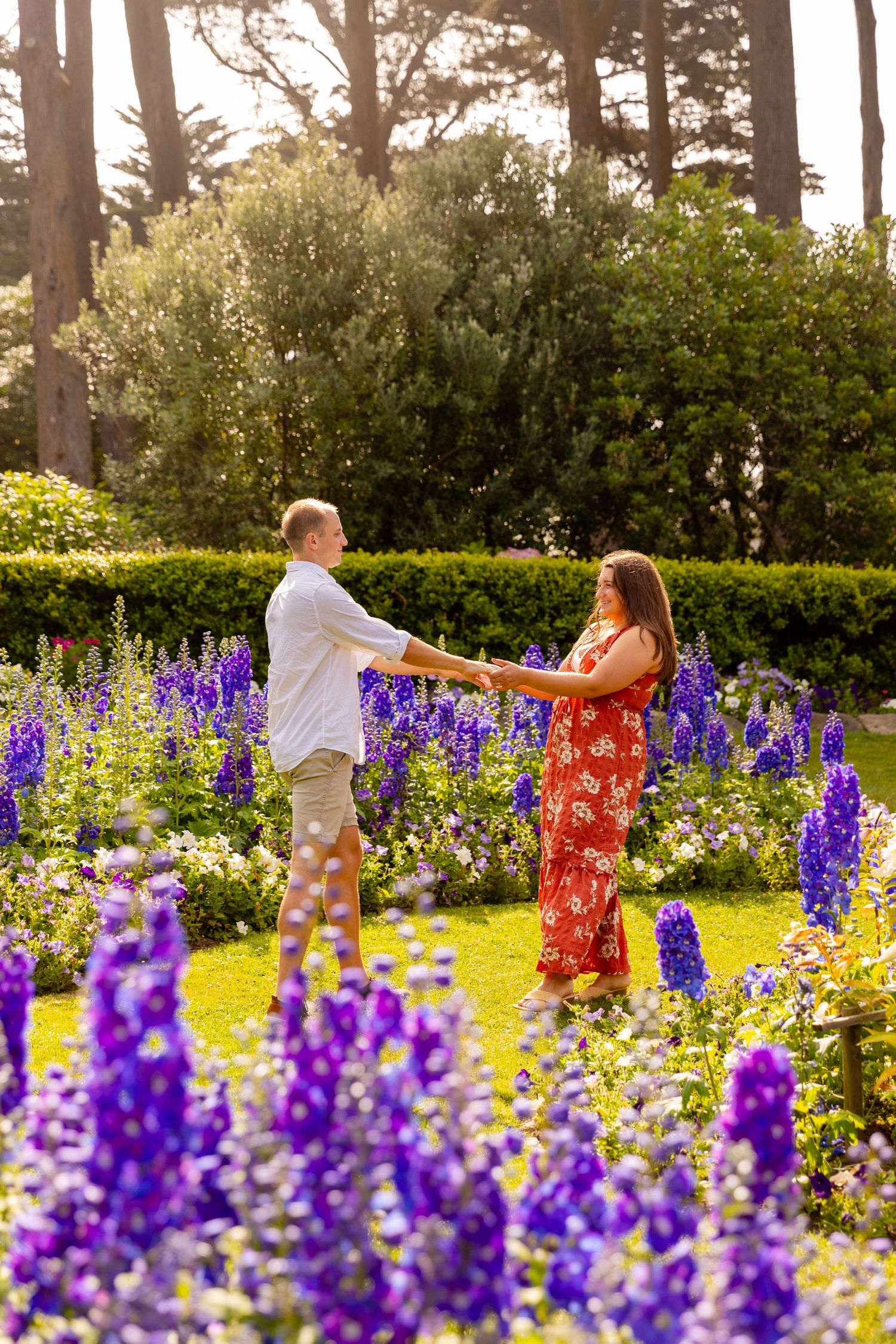 A couple dances together in a garden filled with vibrant purple delphiniums and yellow wildflowers on a sunny day.