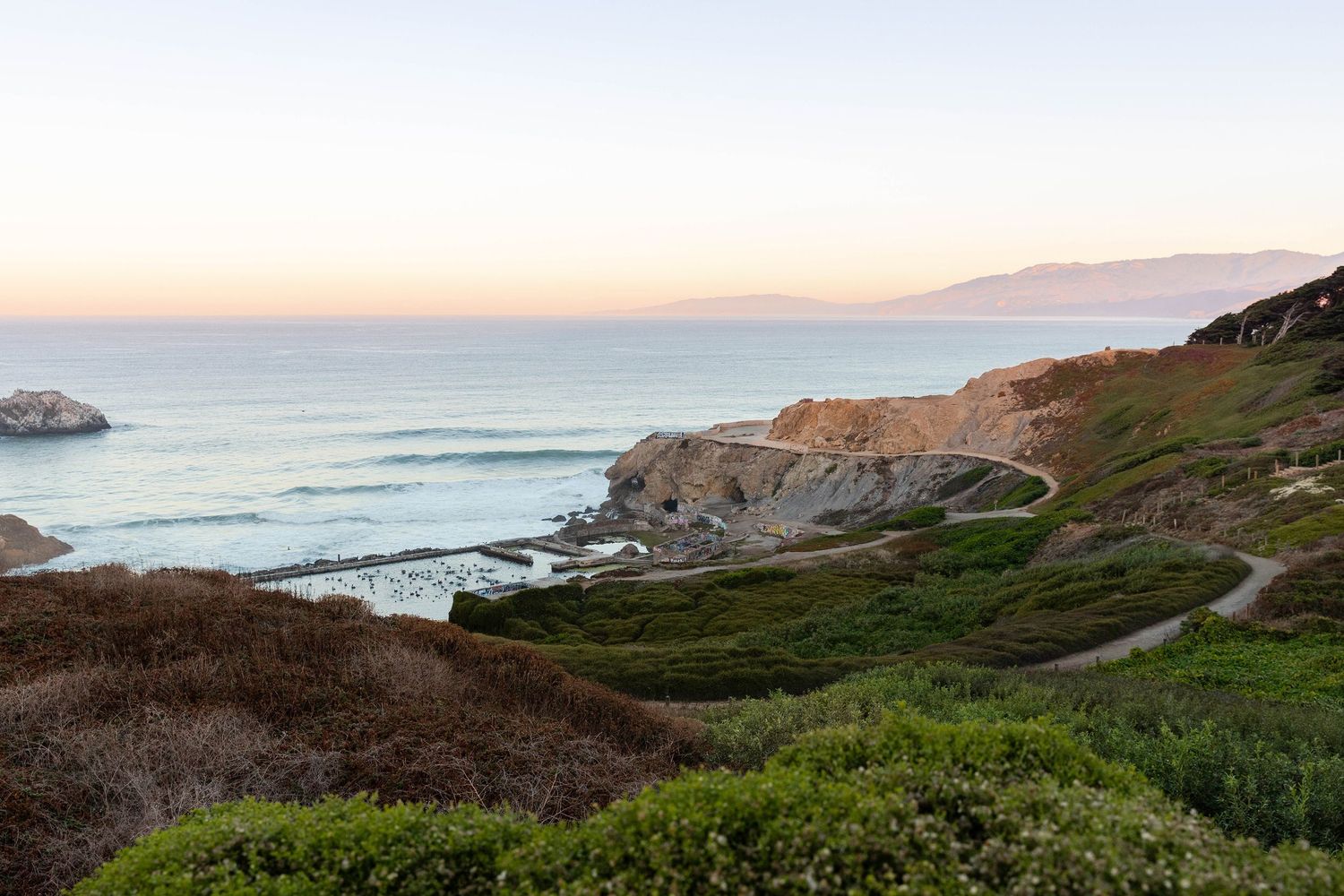 Scenic coastal cliffs with winding path, lush greenery, and ocean waves at sunset along the California coastline.