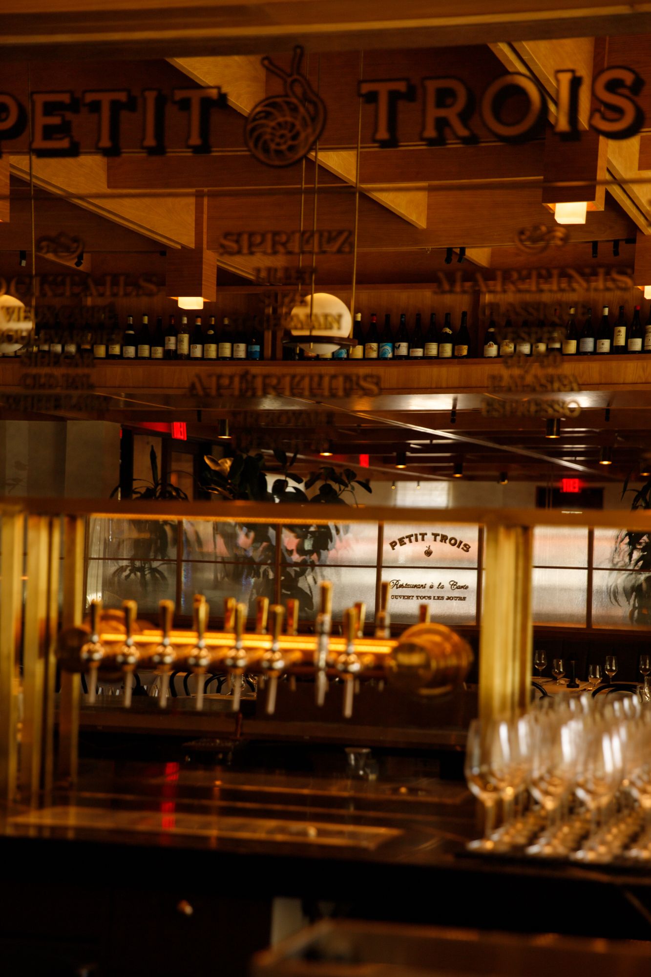 Petit Trois French bistro bar interior with golden beer taps, wine glasses, and bottles displayed on shelves in warm lighting.