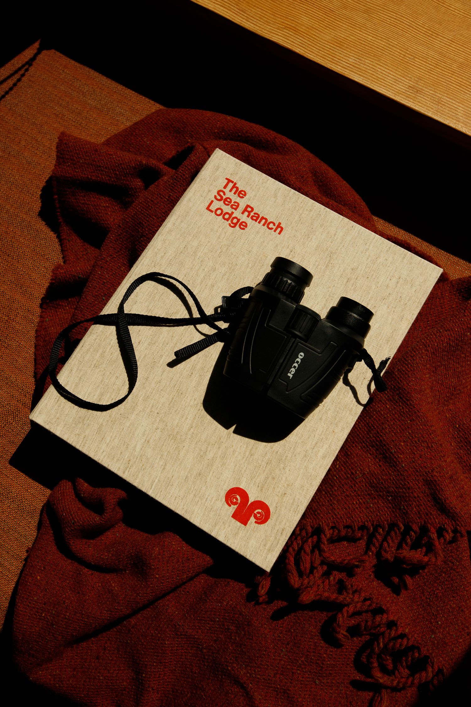 The Sea Ranch Lodge book with black binoculars on a red knit blanket in warm light.