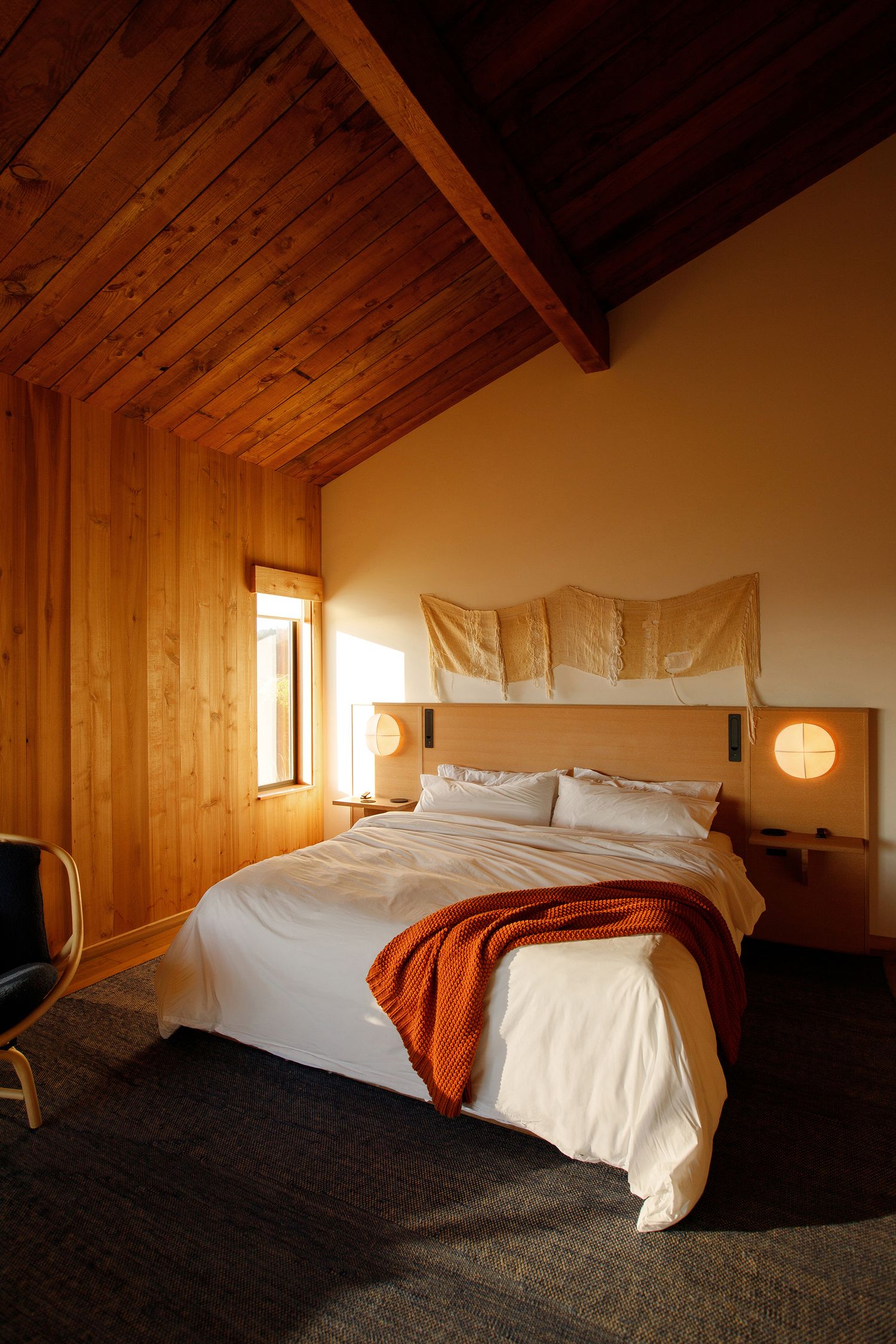 Cozy bedroom with wood-paneled ceiling, white bedding, rust throw blanket, and warm globe sconces flanking a simple headboard.