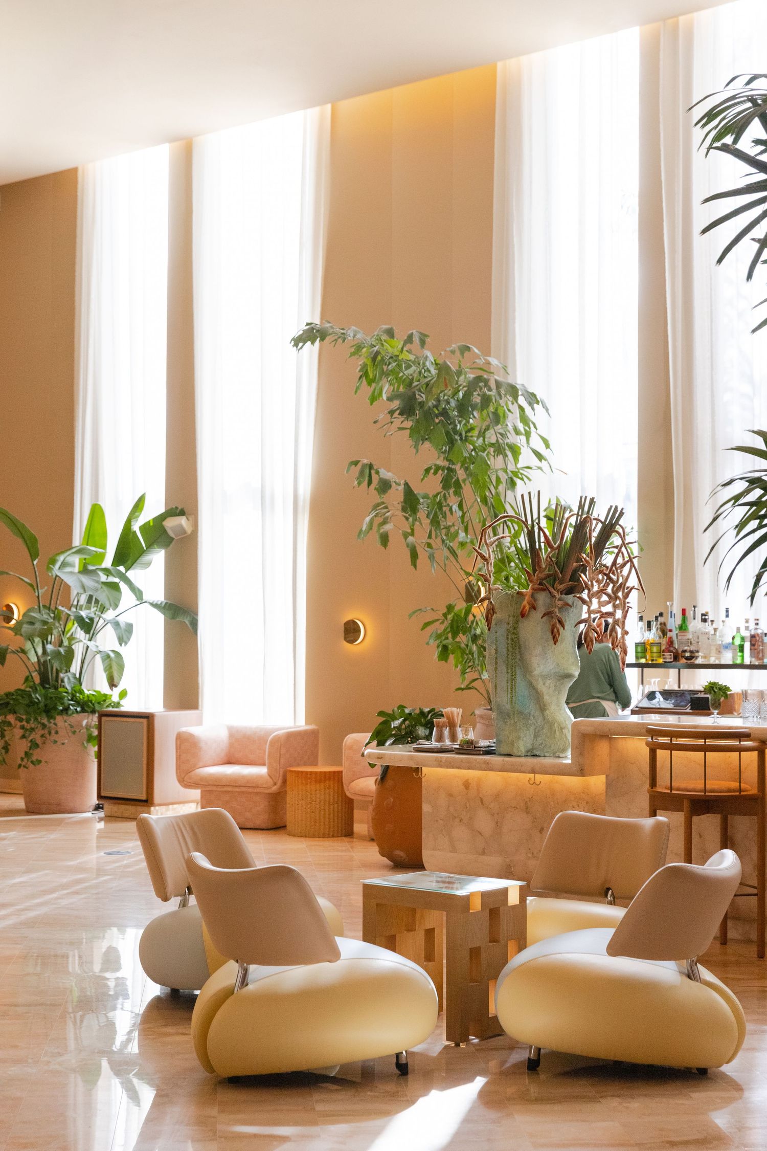 Elegant hotel lobby with cream lounge chairs, marble bar, lush tropical plants, and tall windows with sheer curtains.