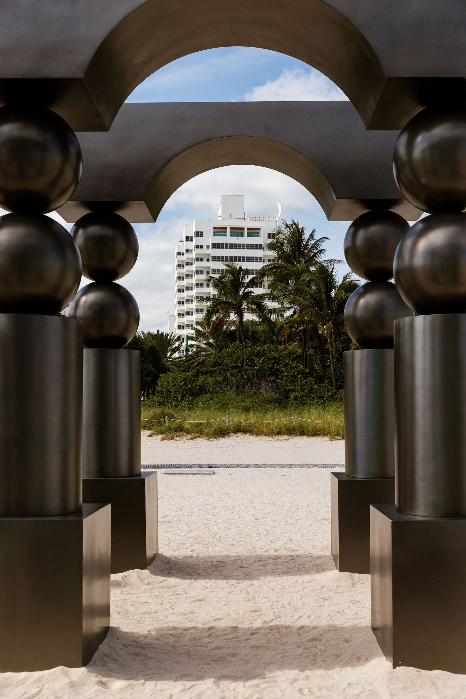 Panoramic view of a sandy beach through metallic architectural columns with palm trees and buildings in the distance.