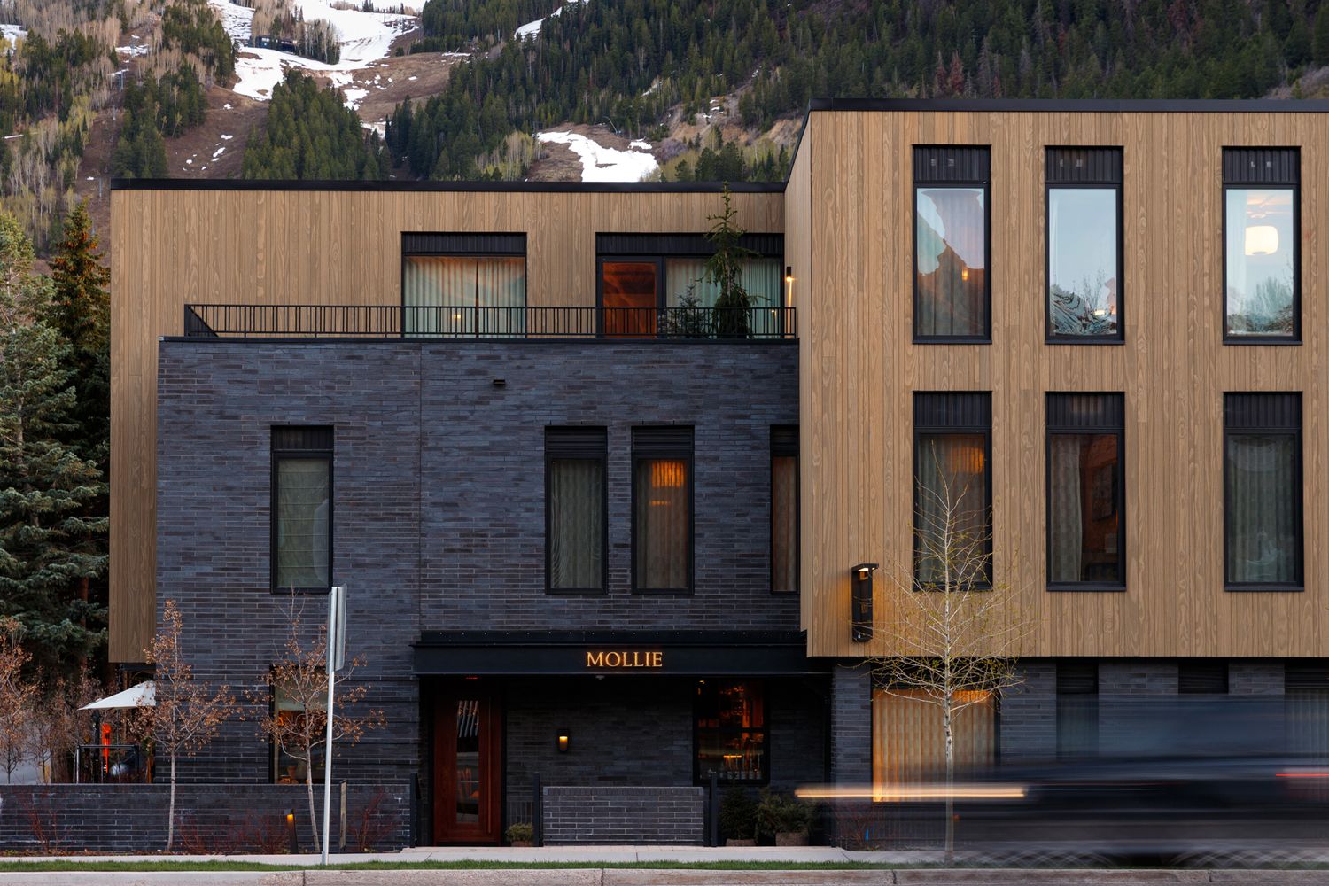 Modern hotel exterior featuring dark stone and wood facade with balcony in a mountain town setting.