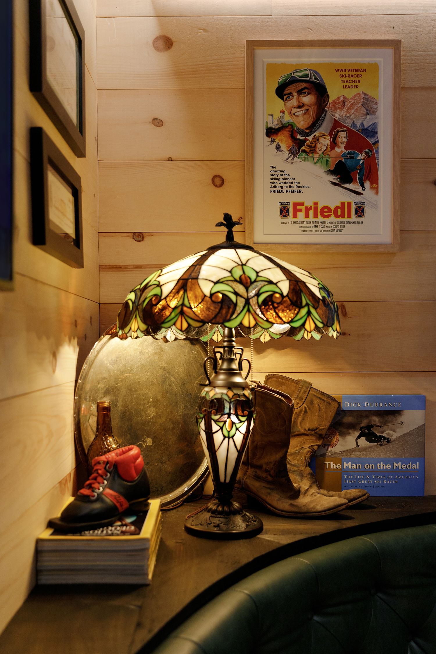 Vintage Tiffany-style table lamp beside ski boots, a Friedl movie poster, and The Man on the Medal book on a wooden shelf.