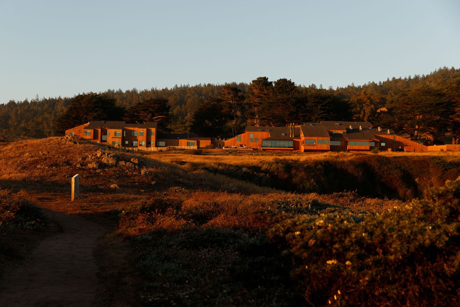 Golden hour light illuminates rustic buildings nestled among trees and golden grasslands in a remote coastal landscape.