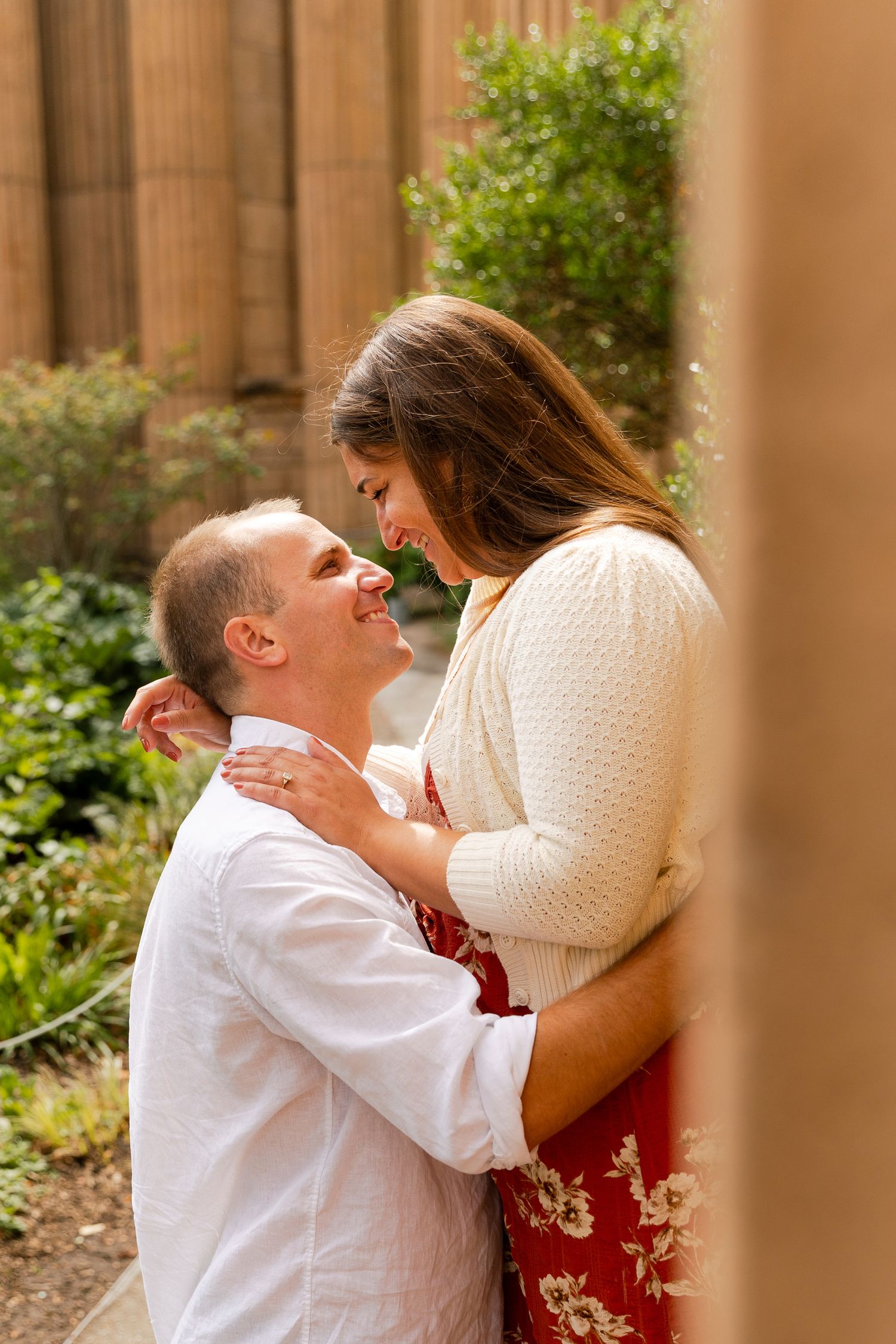 Romantic couple embraces outdoors near stone columns and greenery during an engagement photo session.