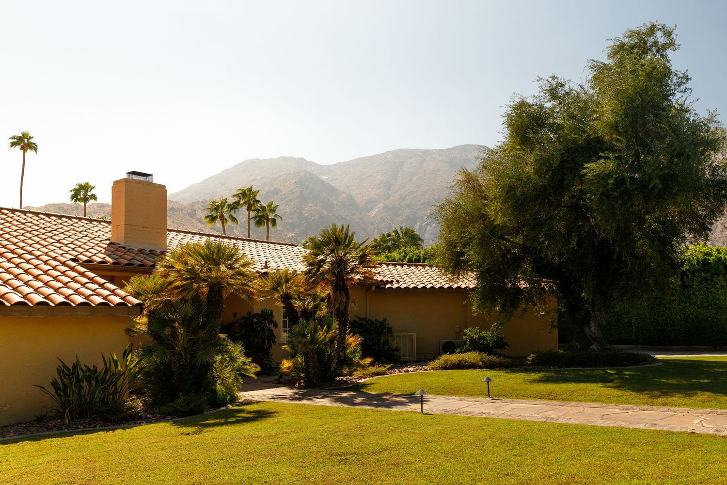 Spanish-style home with red tile roof, palm trees, lush green lawn, and scenic mountain backdrop in Palm Springs.