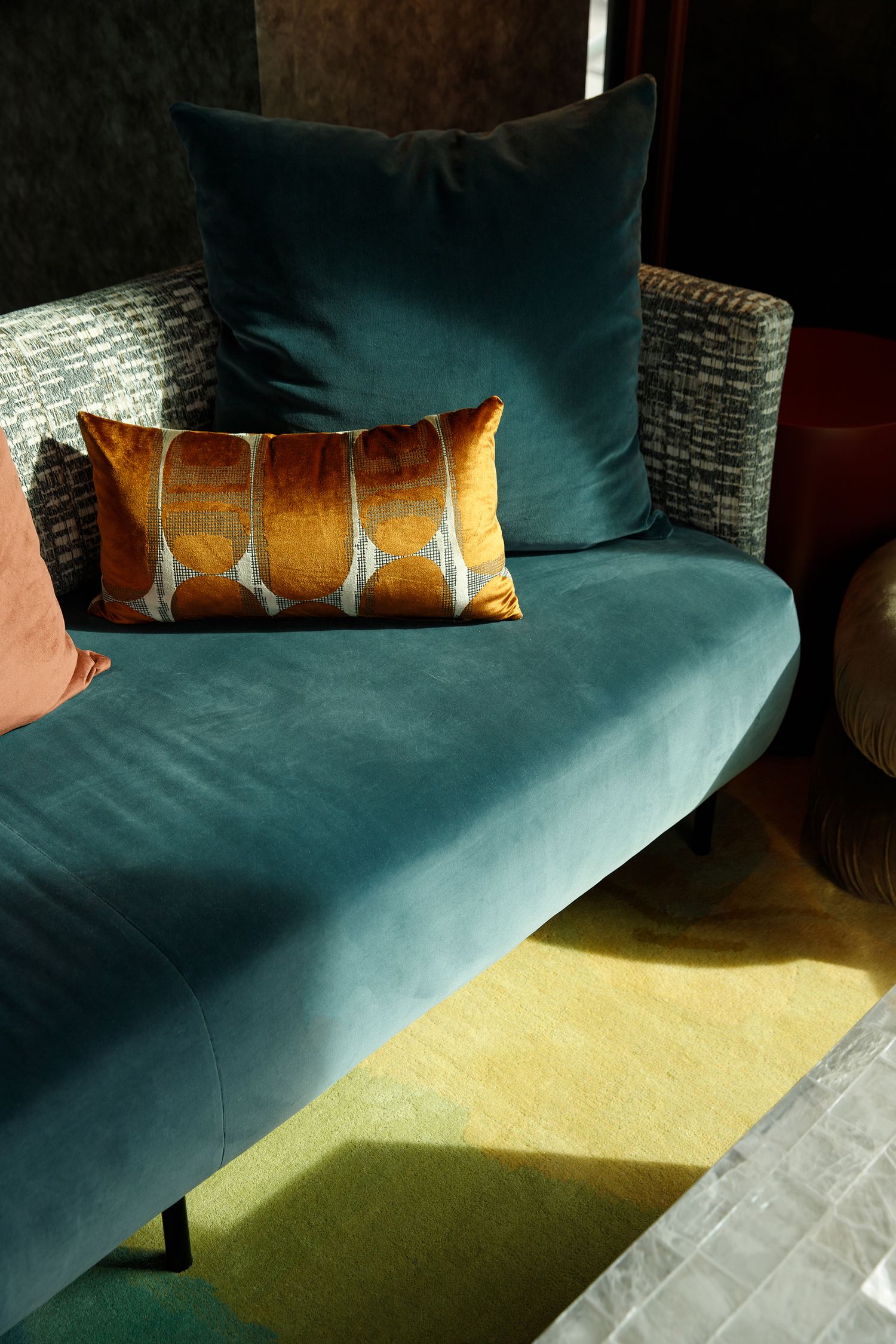 Teal velvet chaise lounge with burnt orange decorative pillow and blue cushion in a stylish modern interior.