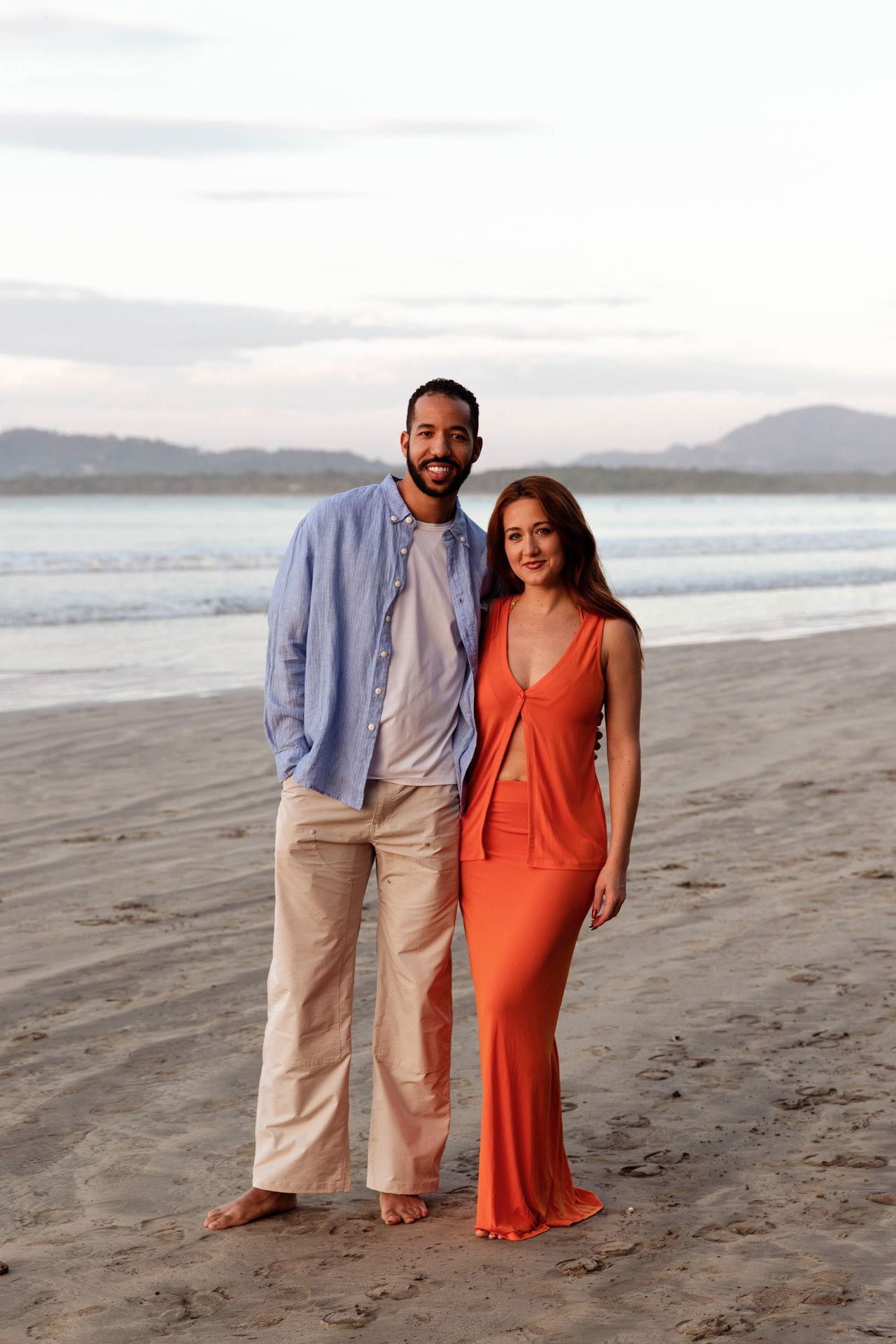 A couple poses together on a scenic beach at sunset with mountains visible in the background.