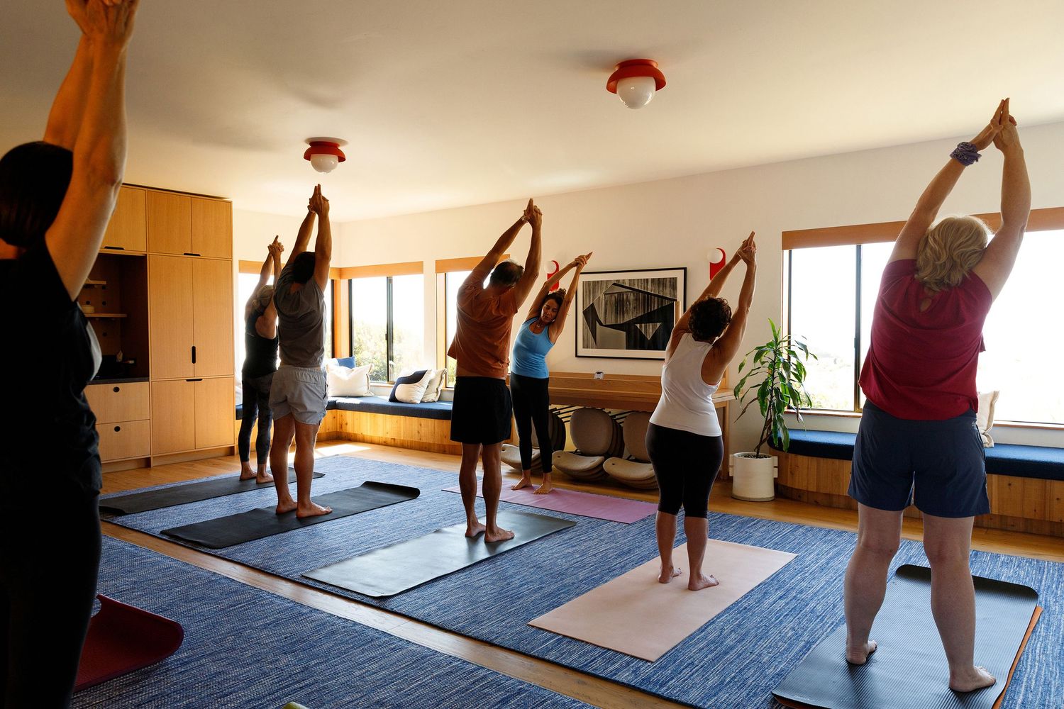 Group yoga class practicing stretching poses on mats in a bright, modern studio with large windows.