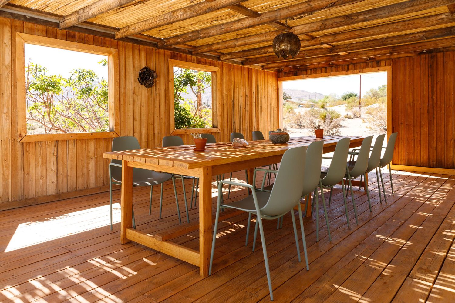 Rustic wooden deck dining area with long table, modern gray chairs, and open desert views under a slatted pergola roof.