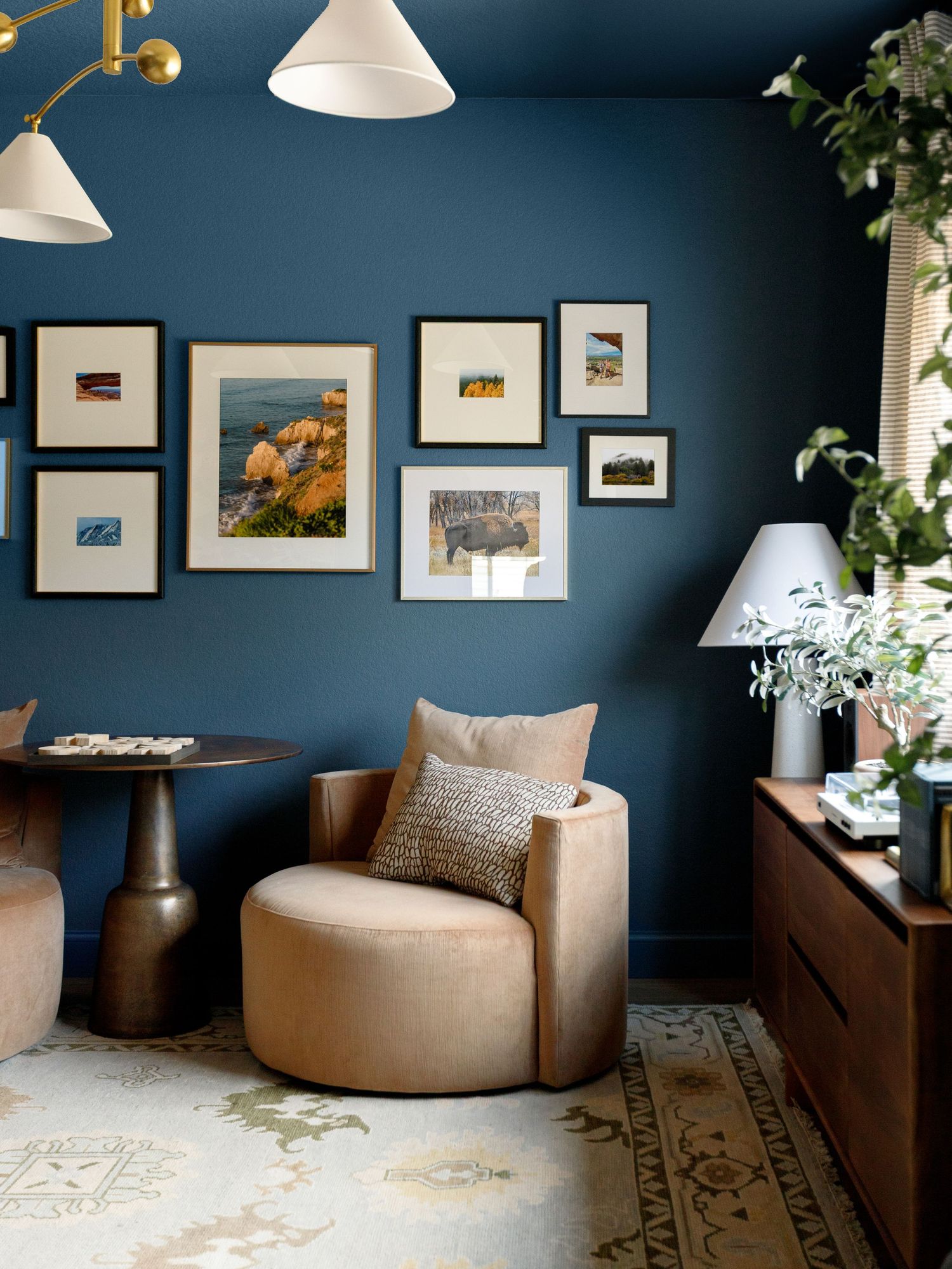 Elegant reading nook with teal blue walls, beige swivel chair, gallery wall art, wood dresser, and modern pendant lights.