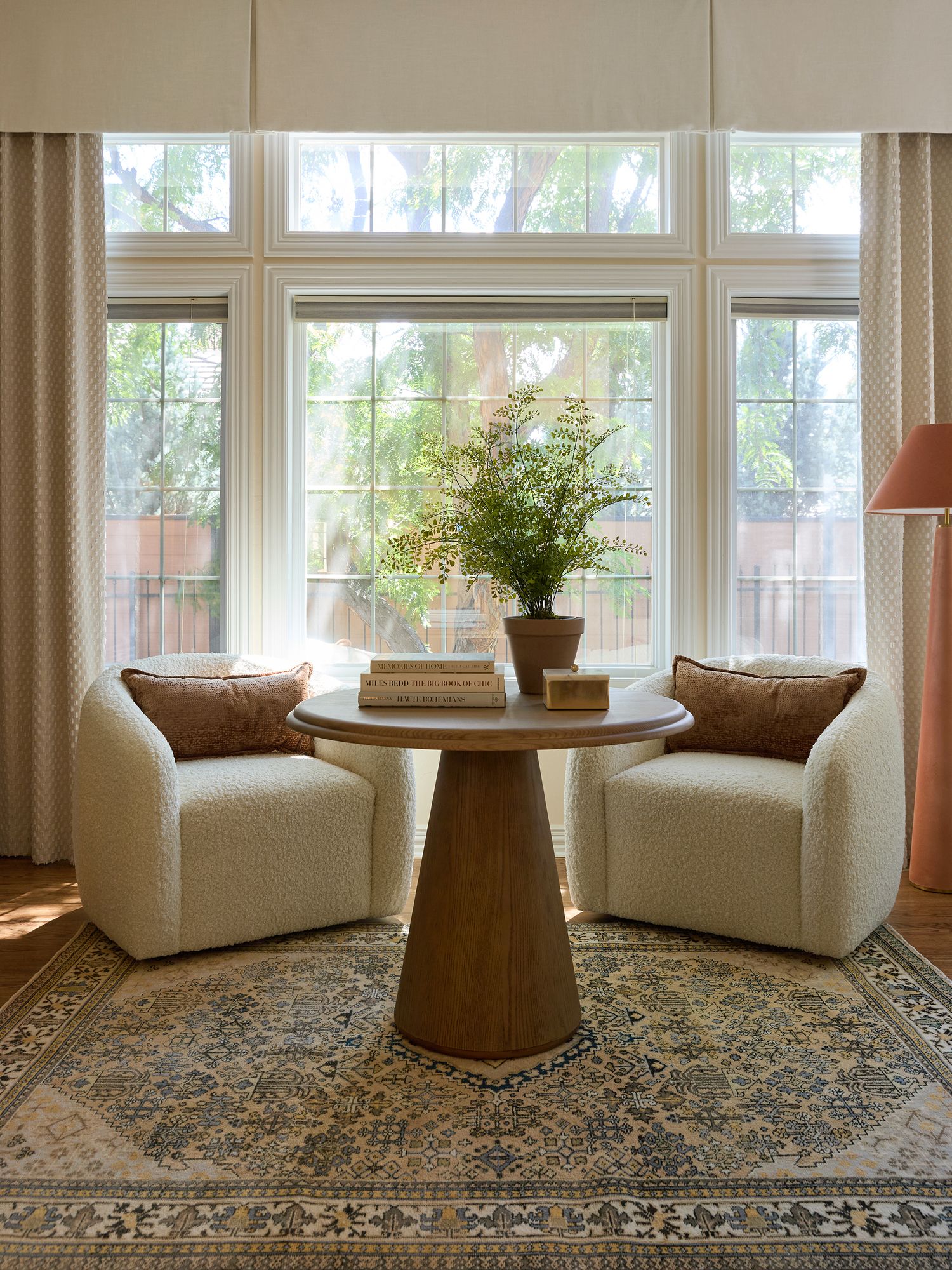 Cozy sitting nook with two white boucle chairs, round wood pedestal table, potted plant, and vintage rug by large windows.