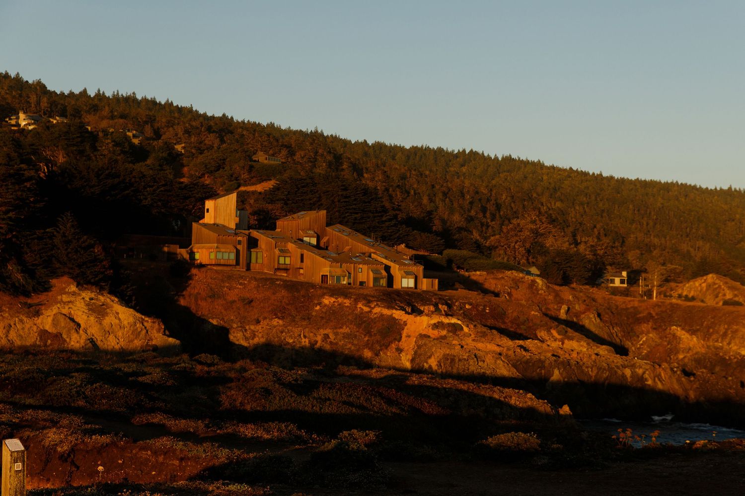 Rustic coastal buildings perched on rocky cliffs at golden sunset with dense evergreen forest backdrop.