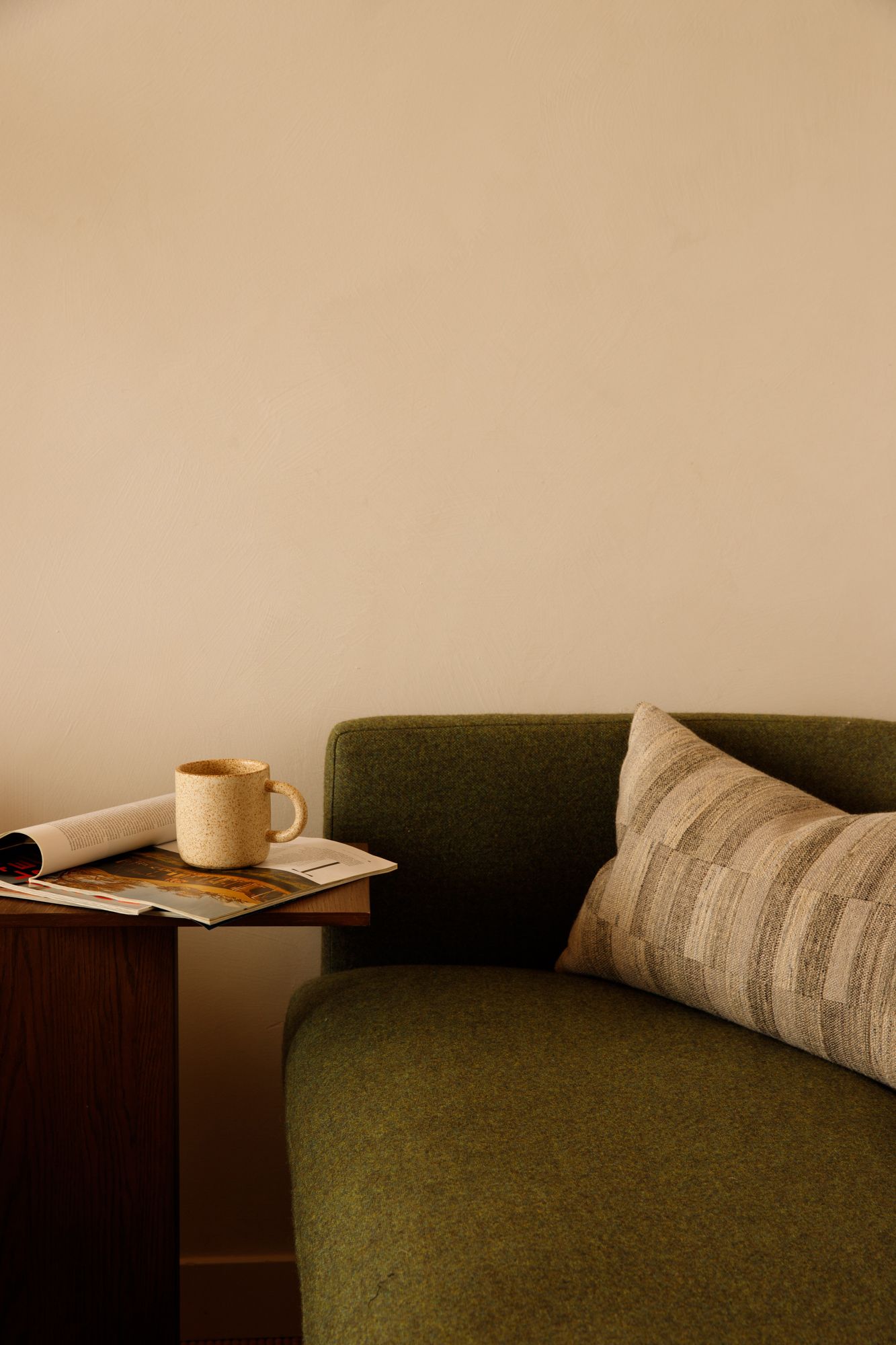 A green wool sofa with a striped lumbar pillow beside a wooden side table holding a ceramic mug and magazine.