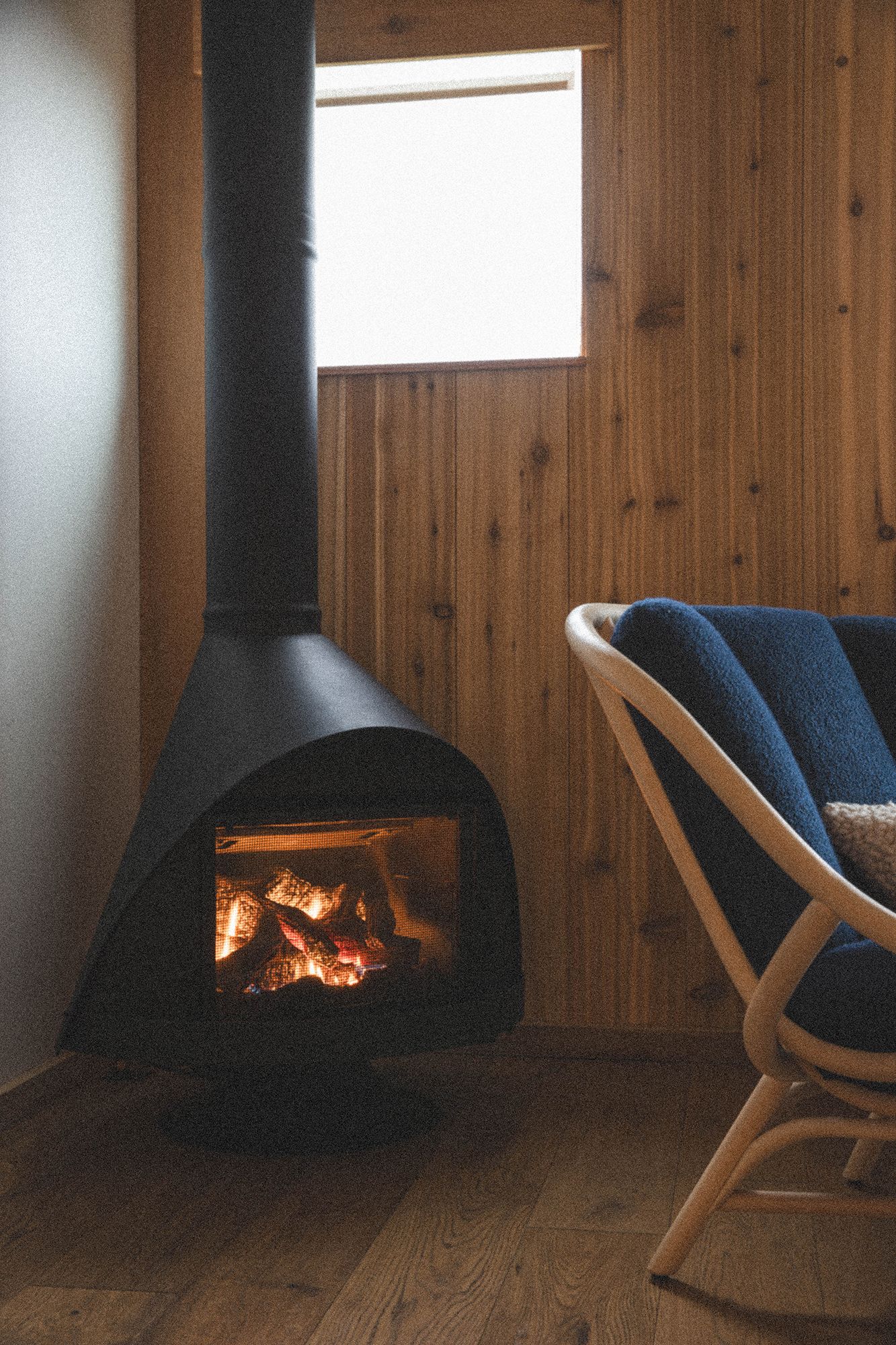 A modern black wood-burning fireplace glows warmly against wooden walls with a rocking chair beside it. Editorial travel imagery.