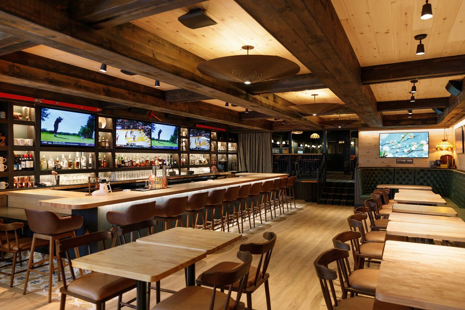 Upscale sports bar with wood beam ceiling, long bar with stools, multiple TVs showing golf, and open dining area.