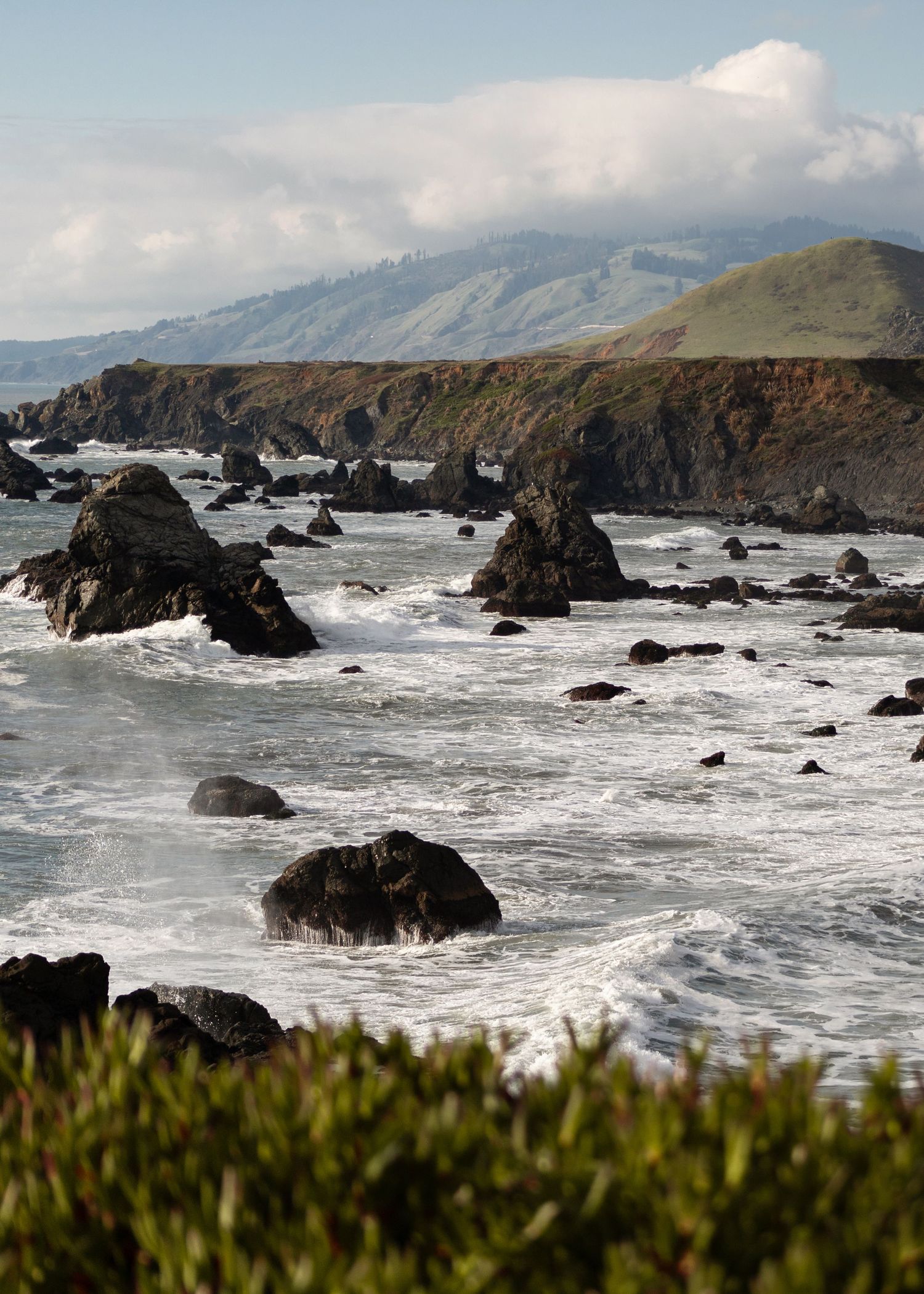 Rocky Pacific coastline with crashing waves, sea stacks, and green hills under a partly cloudy sky.